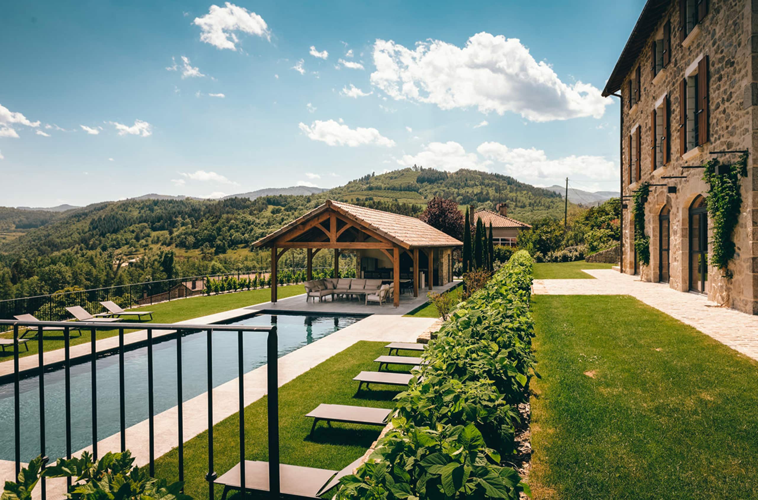 Grand bâtiment en pierre et piscine à débordement dans un jardin paysager, dominant une vallée de collines verdoyantes sous un ciel bleu clair parsemé de nuages.