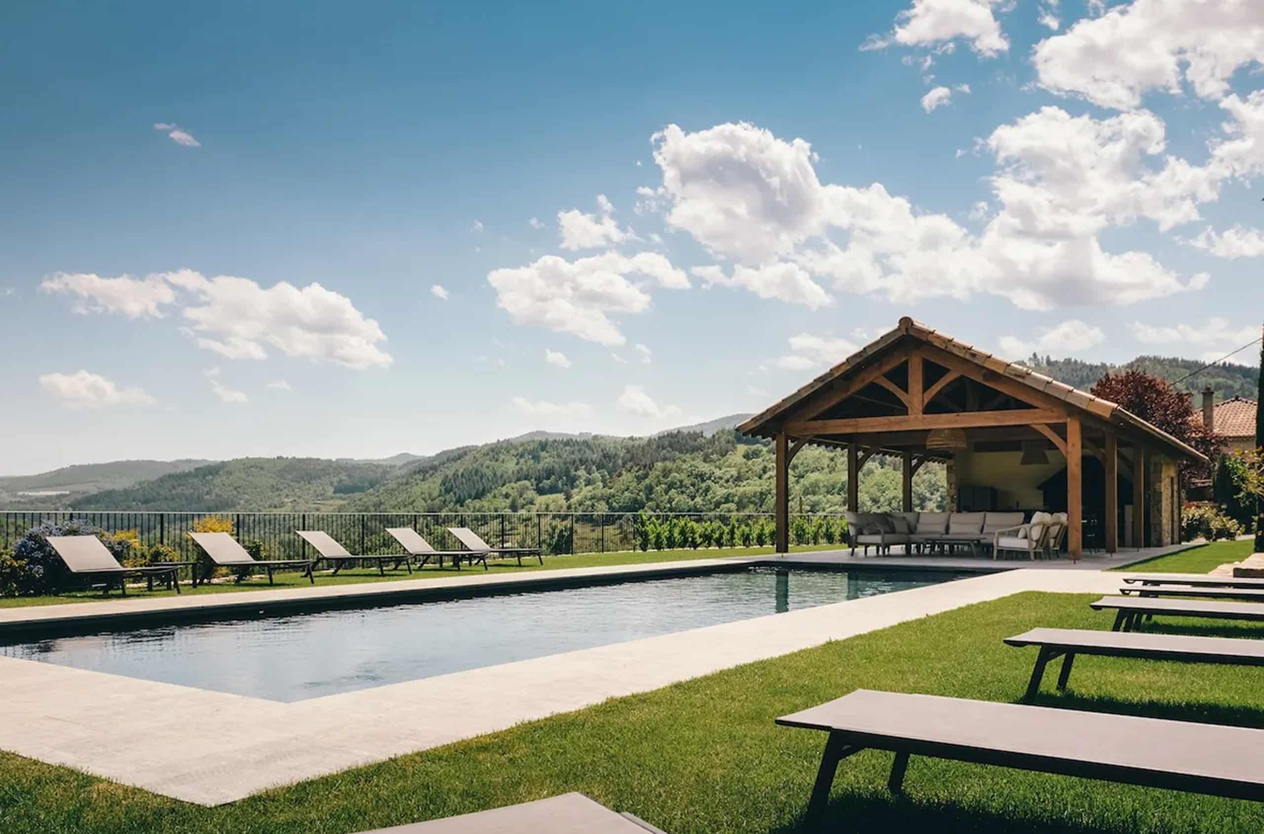 Une piscine rectangulaire bordée de transats sur pelouse fait face à un pavillon. Elle surplombe une vallée montagneuse verdoyante sous ciel bleu nuageux.