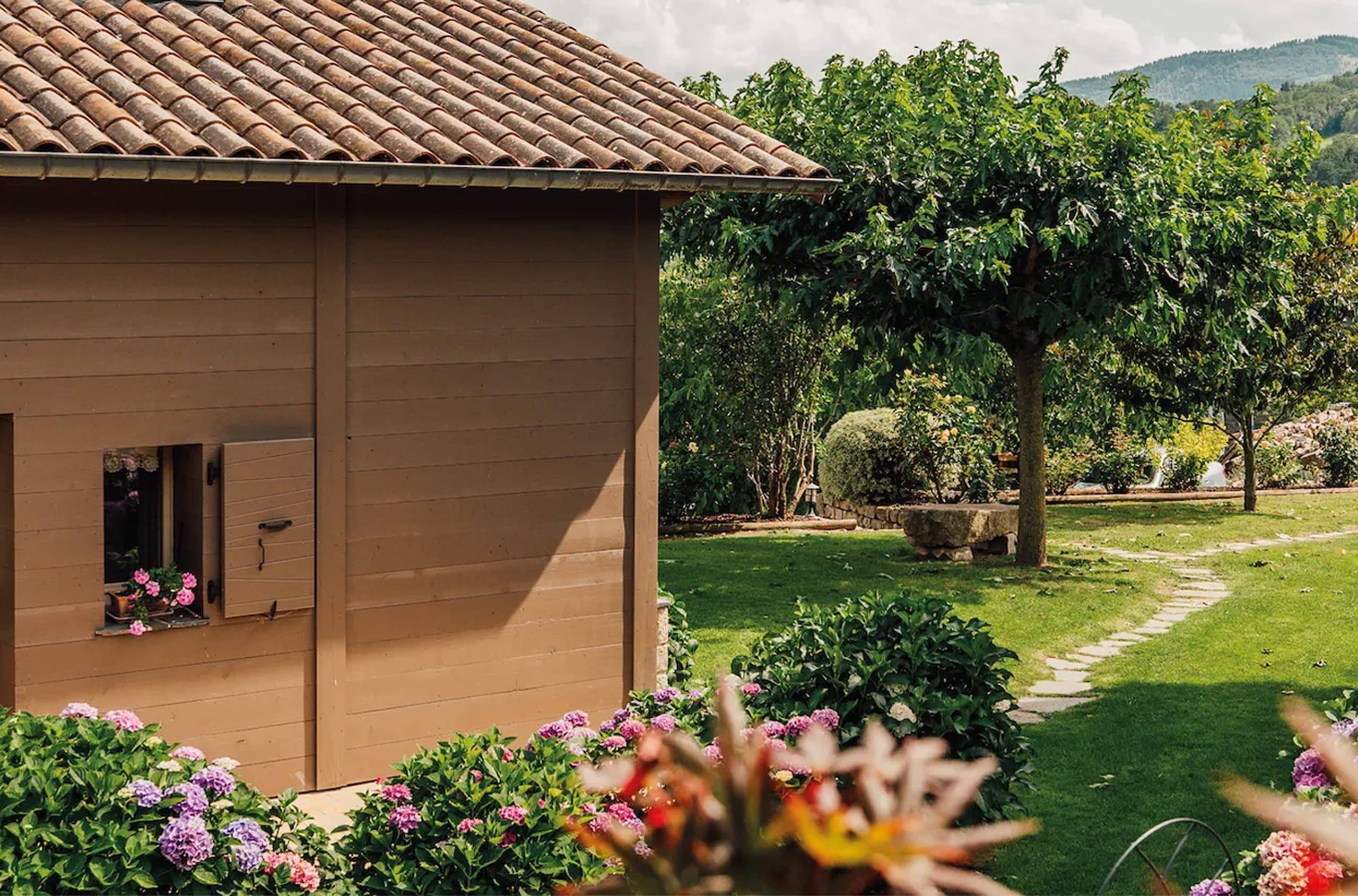 Un bâtiment en bois marron présente un toit de tuiles et une fenêtre fleurie de géraniums roses. Un jardin verdoyant avec arbres, allée dallée et hortensias colorés, sous un ciel nuageux.