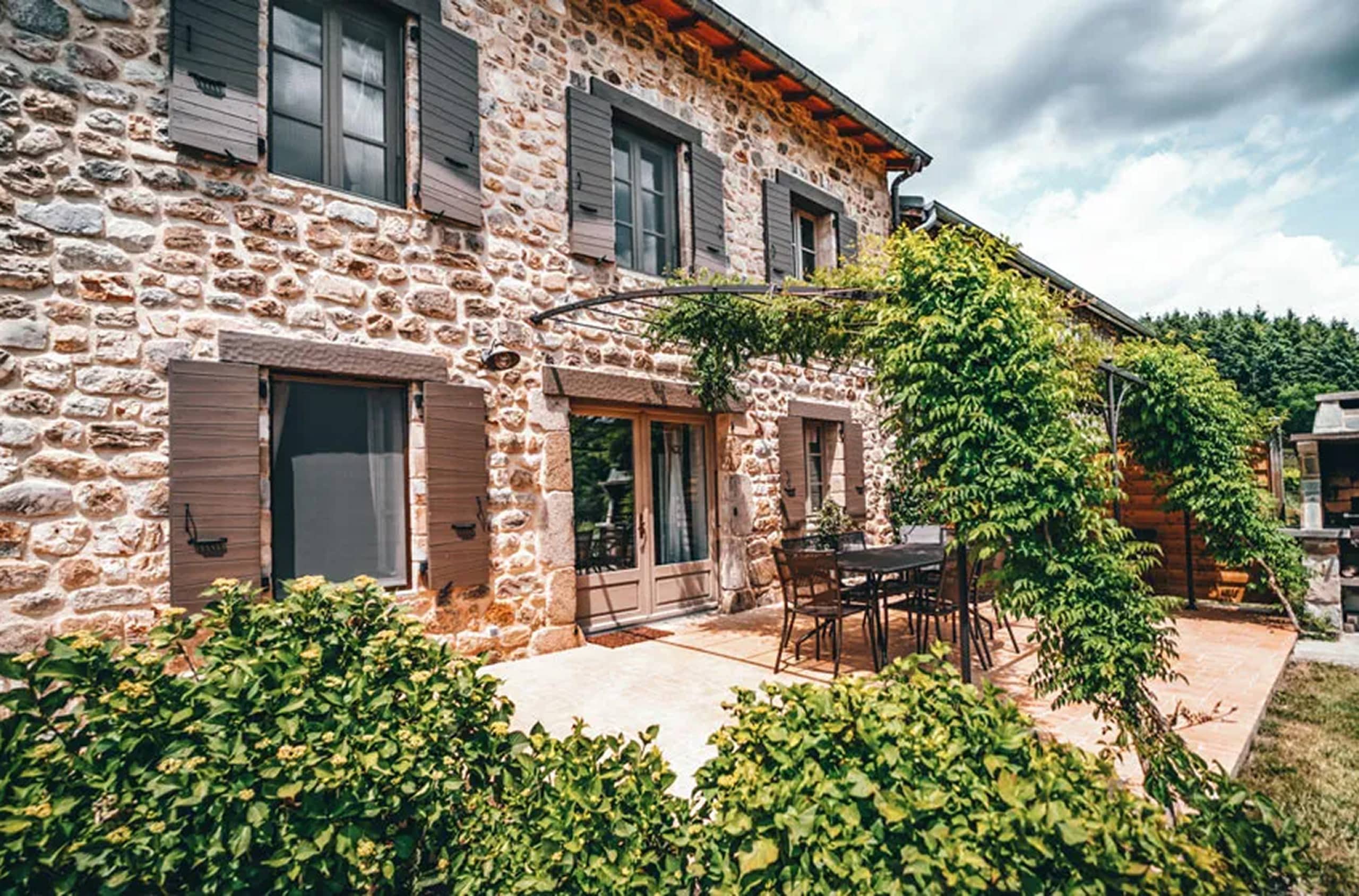 Une maison en pierre présente une façade avec fenêtres à volets gris, un patio meublé et une pergola verdoyante, sous un ciel nuageux, entourée de buissons et d'arbres.
