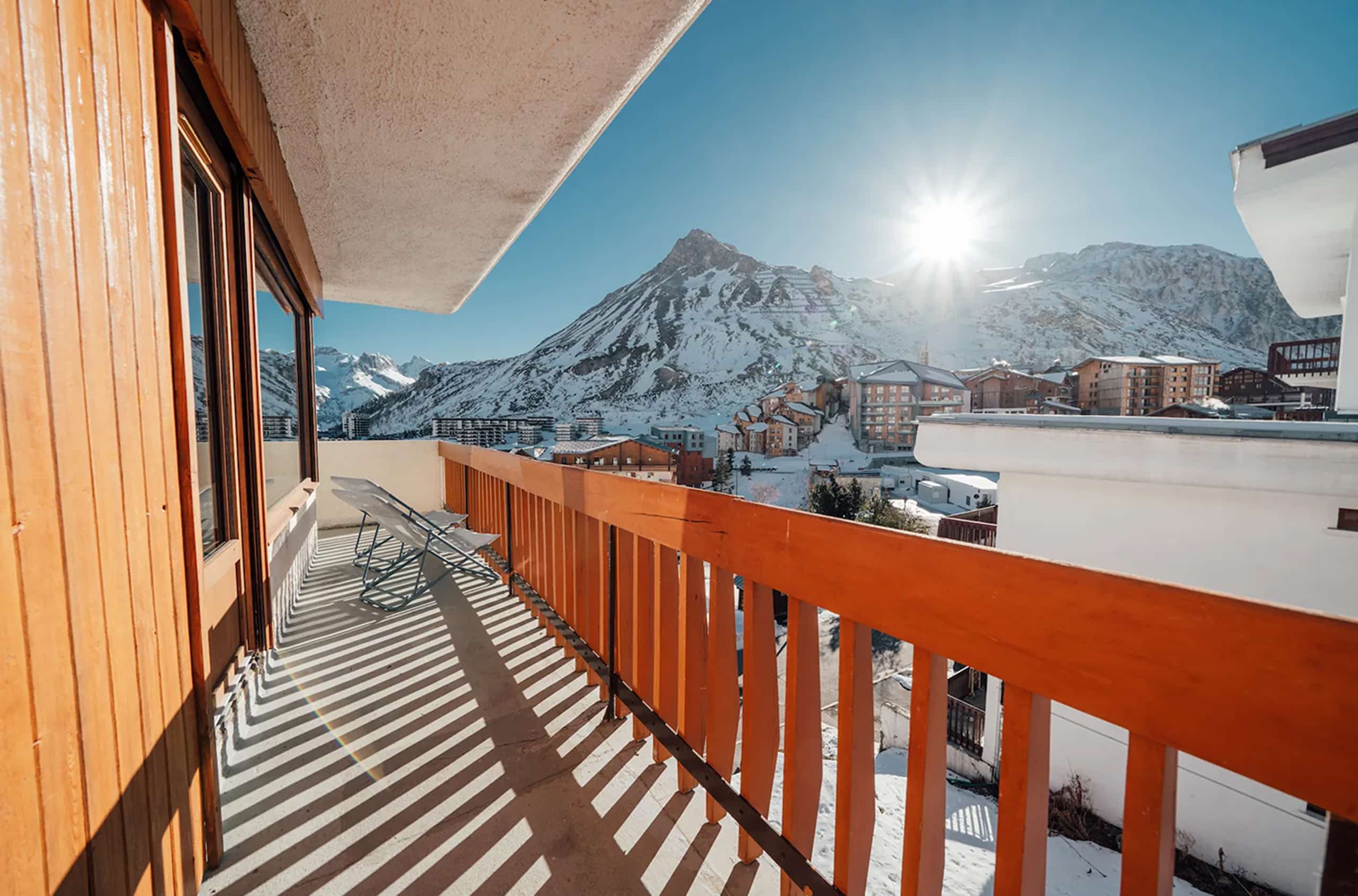 Balcon en bois clair offrant une vue sur un village de ski enneigé. Le soleil hivernal projette de longues ombres sur la terrasse et les montagnes sous un ciel bleu dégagé.