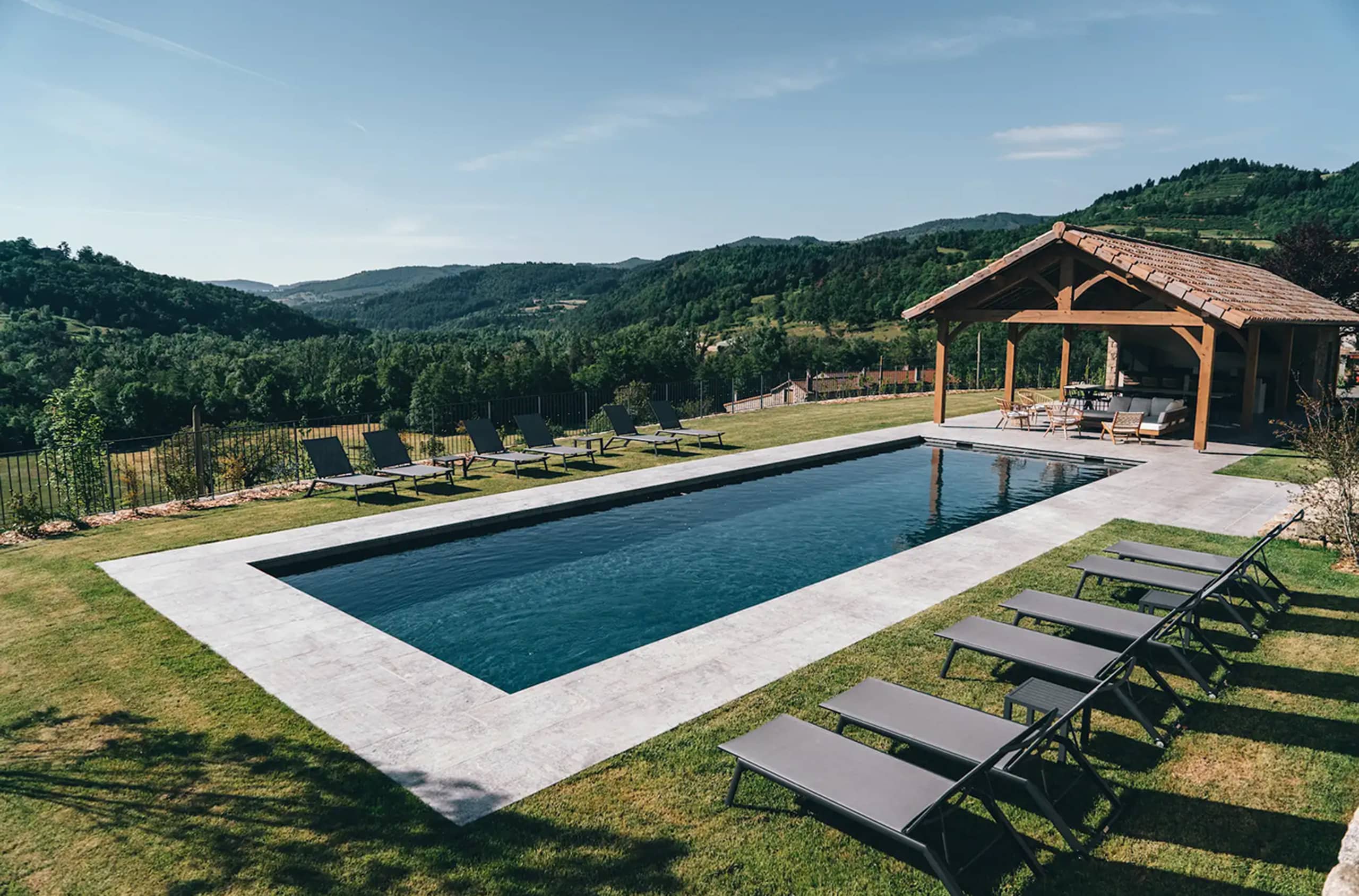 Une longue piscine rectangulaire est bordée de chaises longues et d'un salon couvert, sur une pelouse verdoyante surplombant une vallée et des montagnes boisées sous un ciel clair.