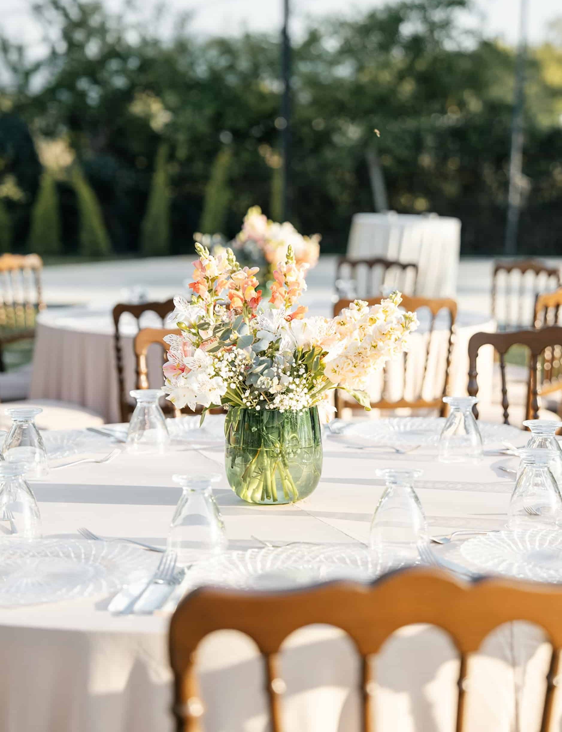 Une table d'extérieur est dressée avec des couverts et un vase vert de fleurs blanches et orangées au centre. Elle est entourée de chaises, d'autres tables et d'arbres flous en arrière-plan.