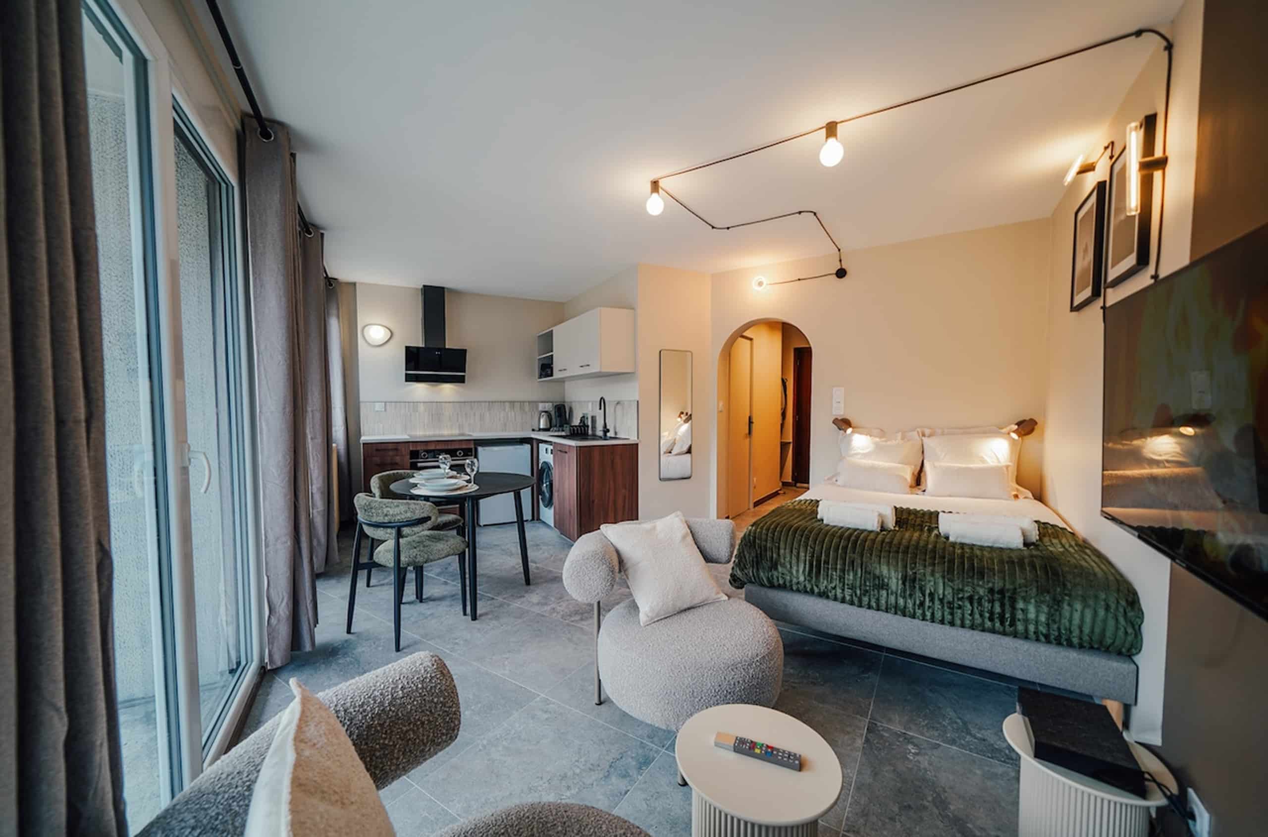 Studio moderne meublé avec lit, cuisine équipée, table à manger et fauteuils. Un grand vitrage éclaire cet intérieur contemporain aux tons neutres et au sol carrelé gris.