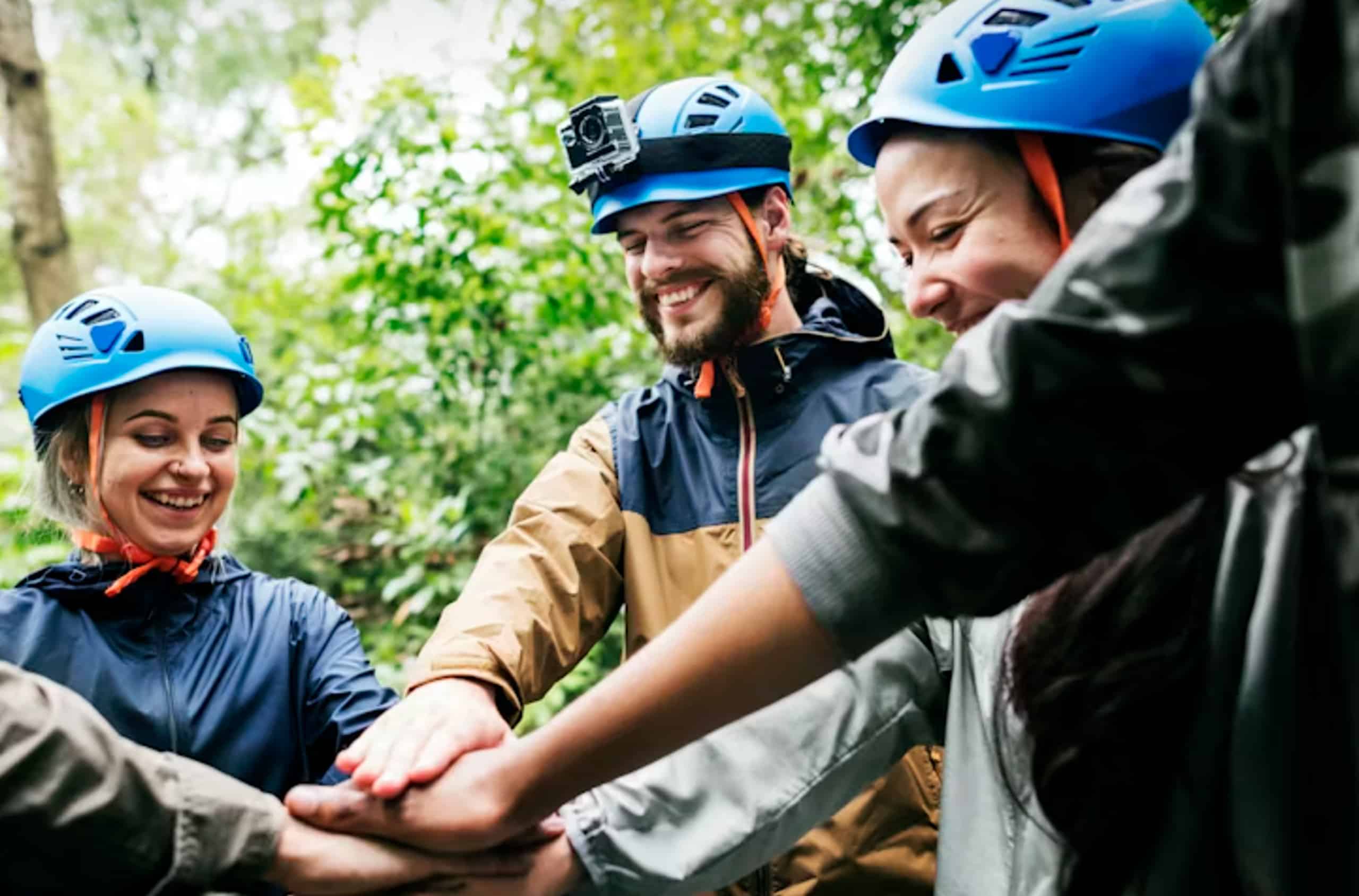 Des jeunes adultes souriants, portant des casques bleus, superposent leurs mains en signe d'unité lors d'une activité d'aventure en pleine nature.