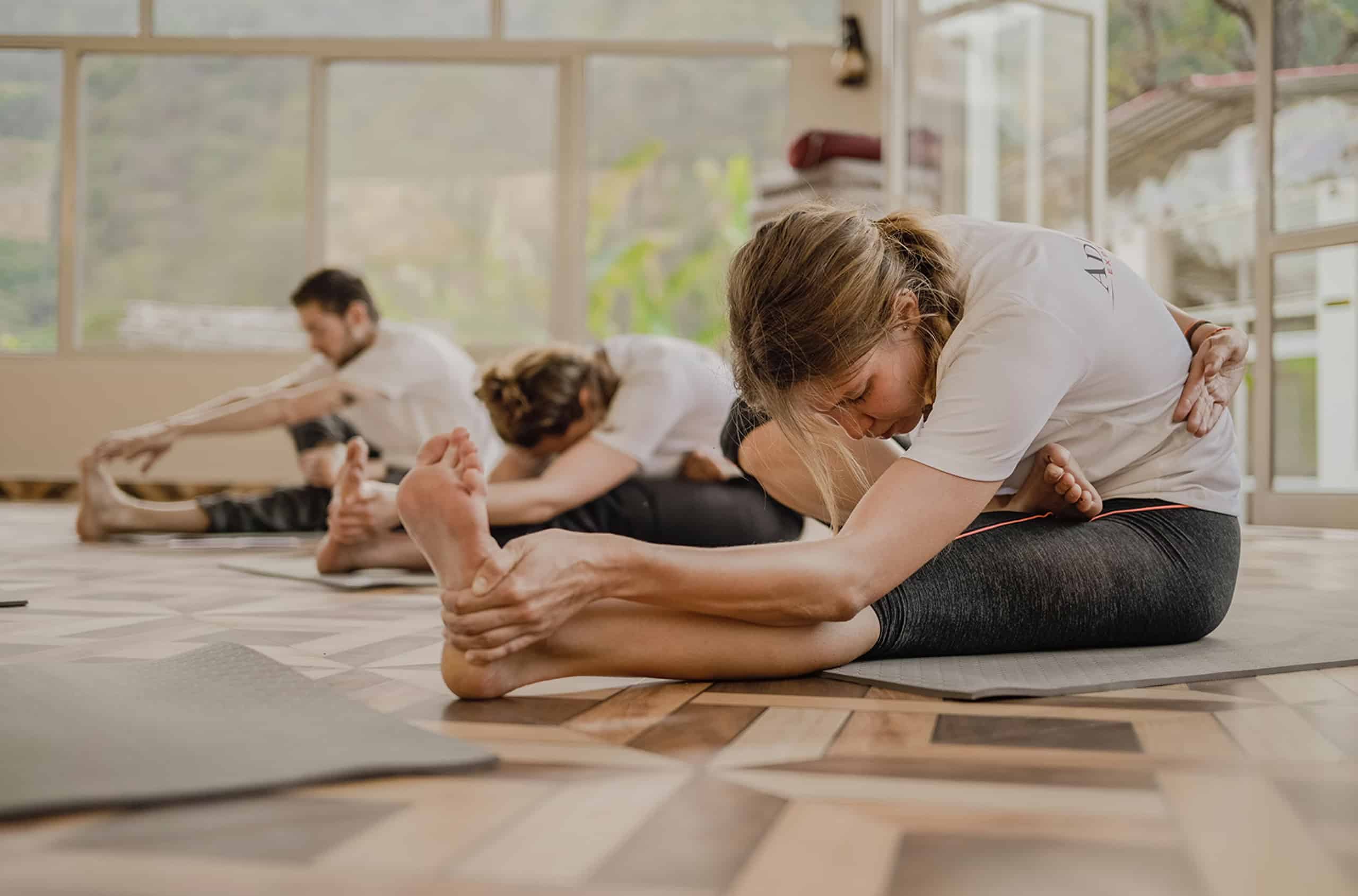 Trois personnes s'étirent vers l'avant, assises sur des tapis de yoga, dans une salle lumineuse. La femme au premier plan, en t-shirt blanc "AD", tient son pied.