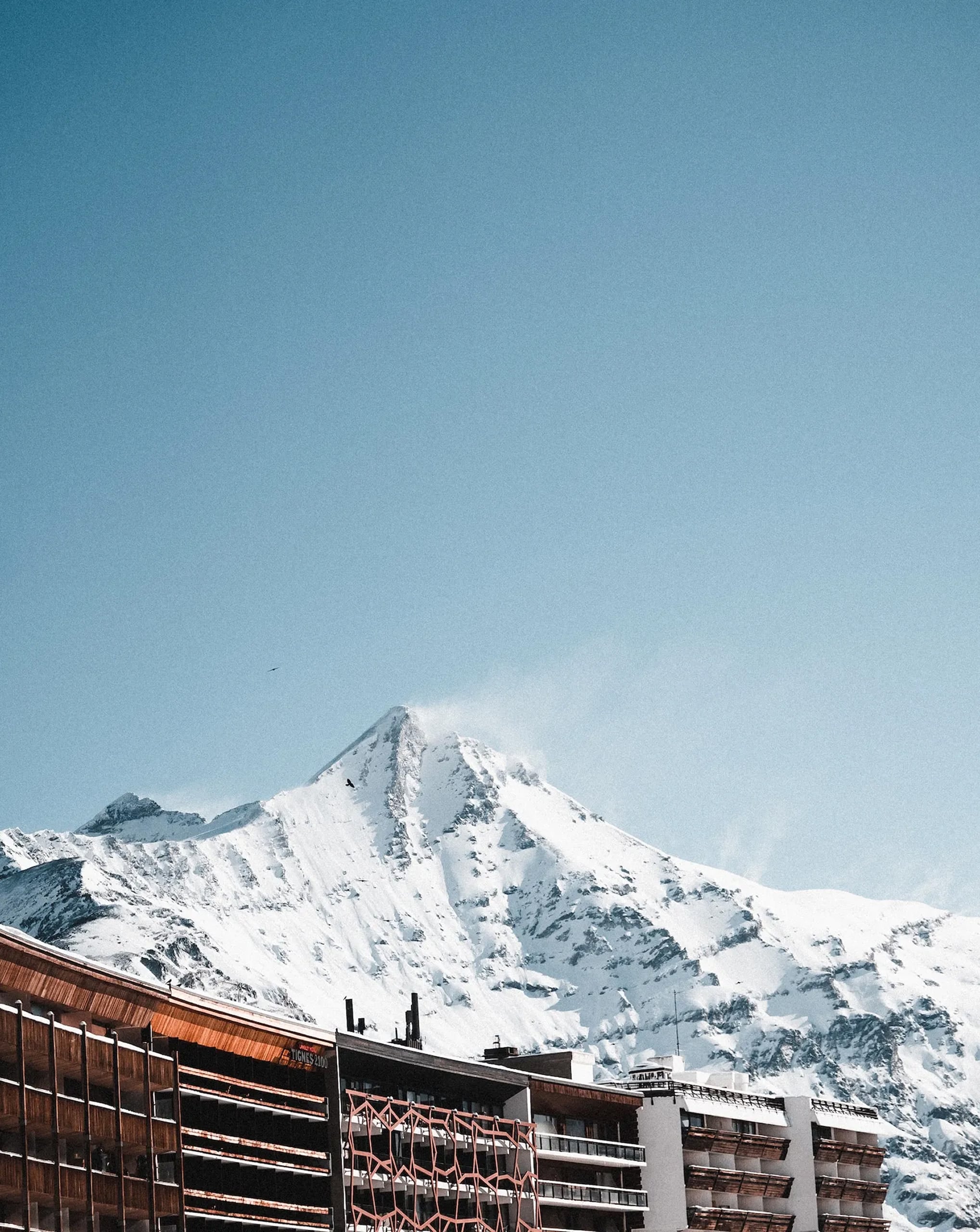 Un pic montagneux enneigé s'élève sous un ciel bleu et pur, des brumes légères flottant à son sommet. Au premier plan, des résidences alpines modernes avec balcons s'étendent. Texte : "WELCOME TIGNES 2100".
