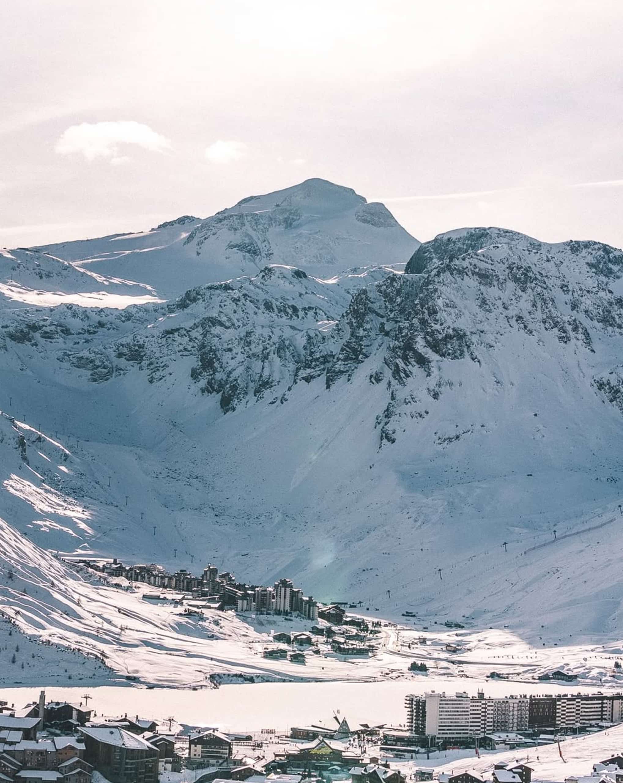 Village de montagne et bâtiments de station de ski dans une vallée enneigée, avec un lac gelé, entouré de vastes pentes skiables et de montagnes imposantes. Texte visible sur un bâtiment : "ETIGNES".