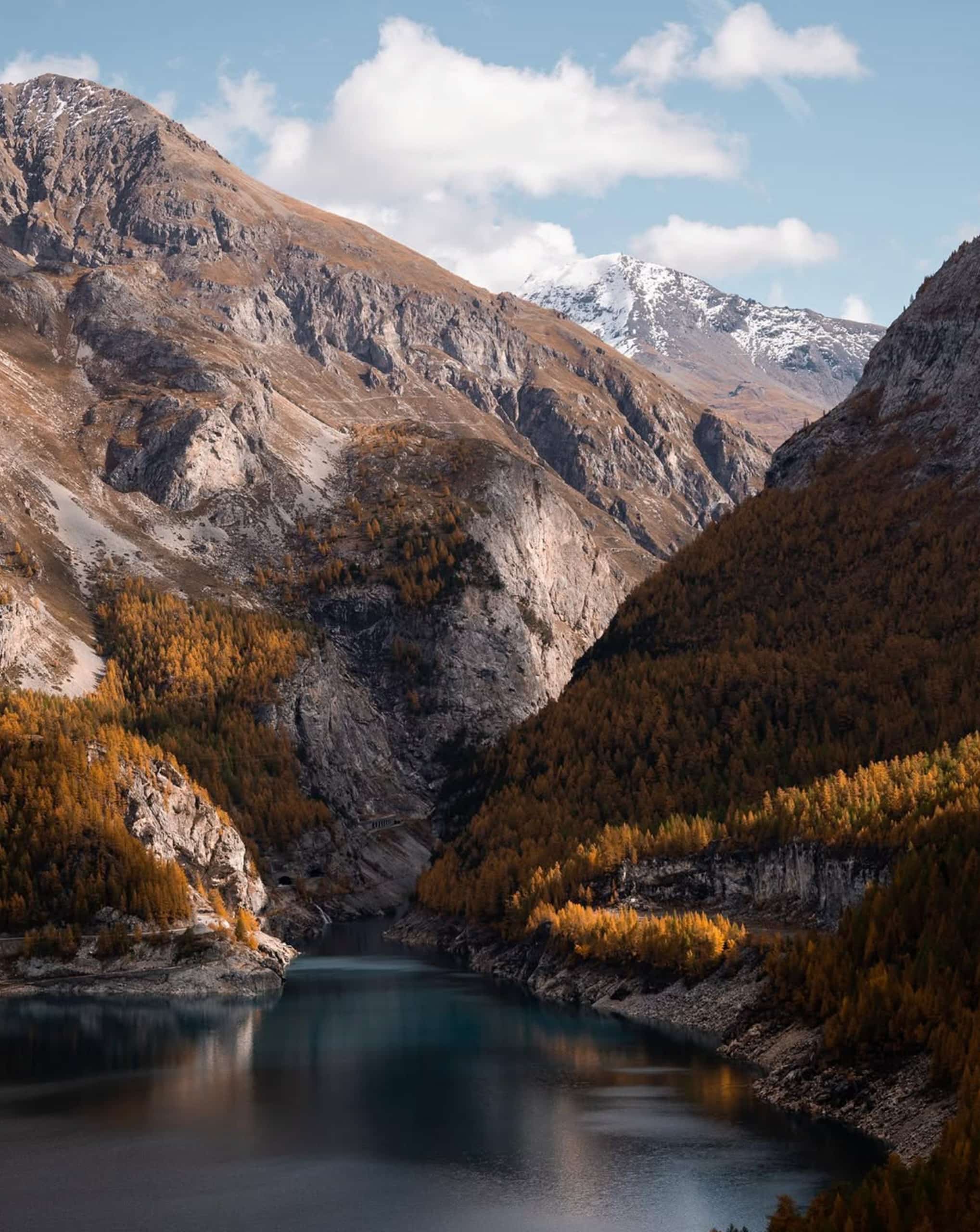 Lac de montagne aux eaux sombres s'étirant entre de hautes montagnes escarpées. Les pentes sont couvertes de forêts dorées d'automne, avec des sommets enneigés sous un ciel nuageux.