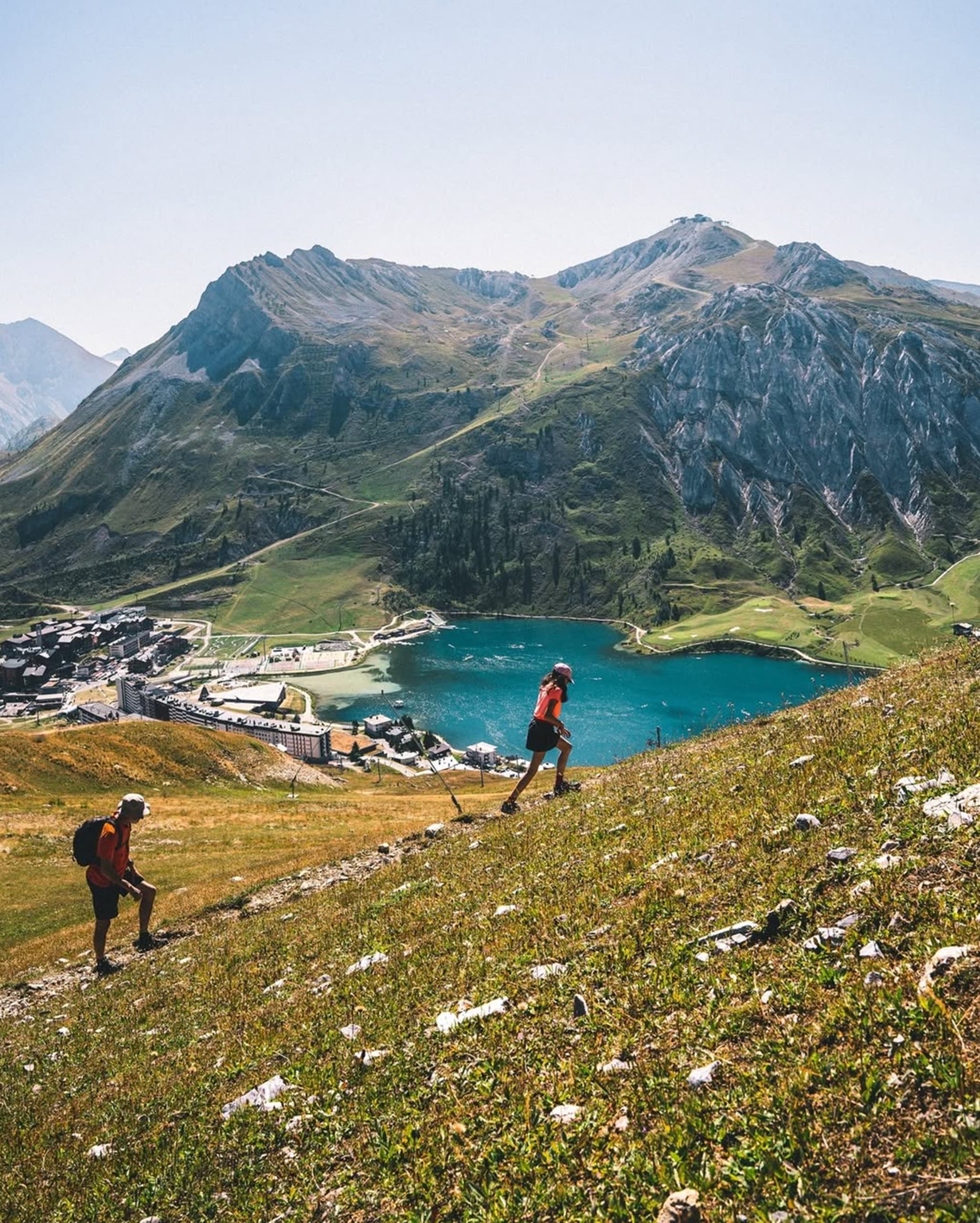 Deux randonneurs montent une pente herbeuse. En arrière-plan, un lac turquoise, un village et des montagnes imposantes s'étendent sous un ciel clair.