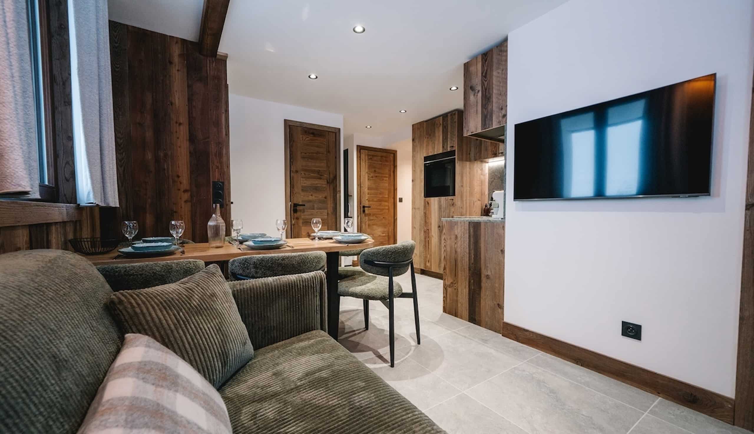 Un salon/salle à manger moderne est aménagé avec une table dressée pour quatre, un canapé vert et une kitchenette intégrée. Des accents boisés réchauffent l'espace, avec une télévision murale.