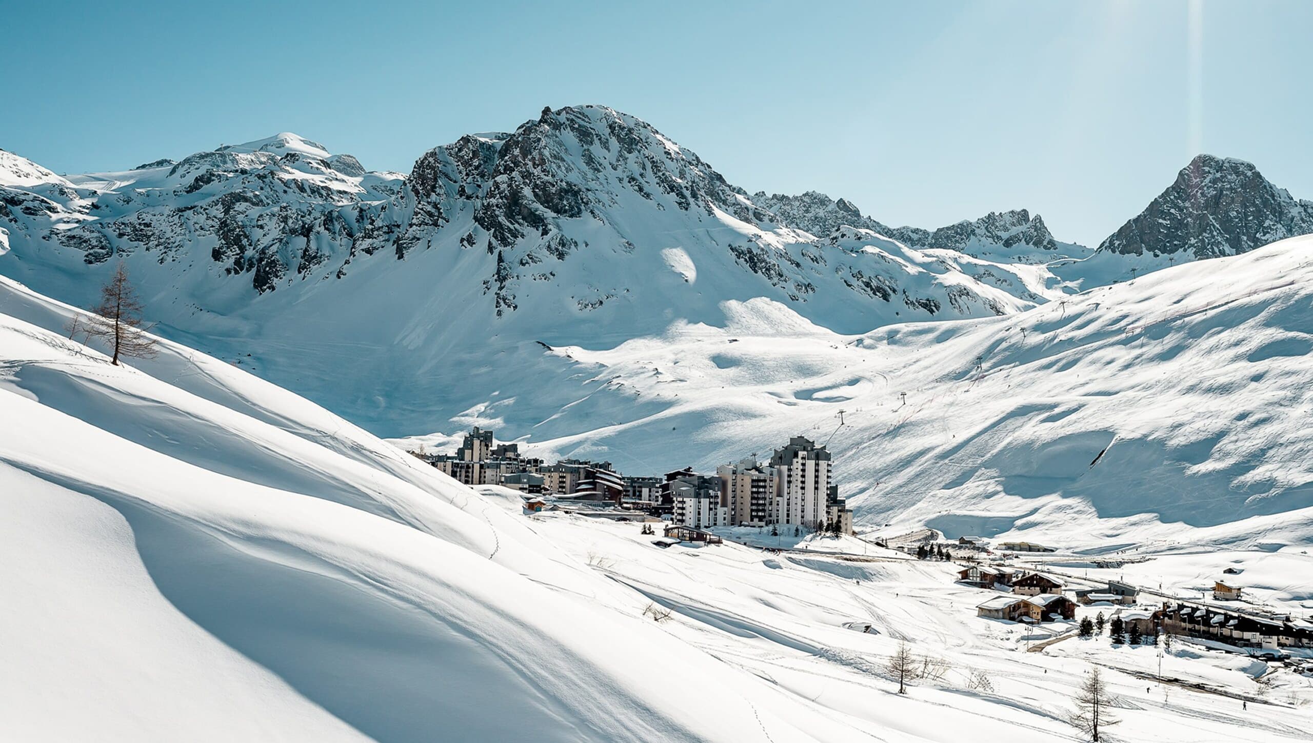 Des bâtiments de station de ski modernes et des chalets sont établis dans une vaste vallée enneigée, dominée par d'imposantes montagnes sous un ciel d'azur.