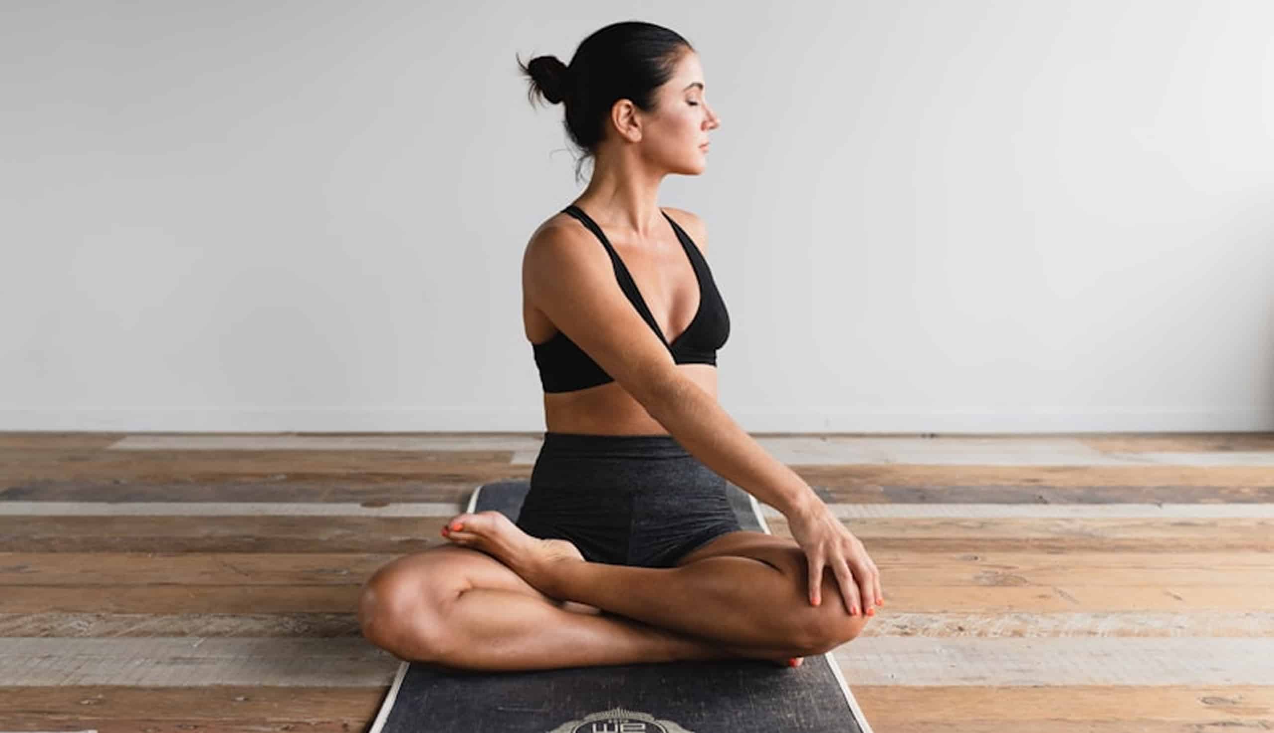 Femme assise en tailleur sur un tapis de yoga, le torse légèrement tourné, yeux clos, pratiquant le yoga ou la méditation dans une pièce épurée au sol en bois.