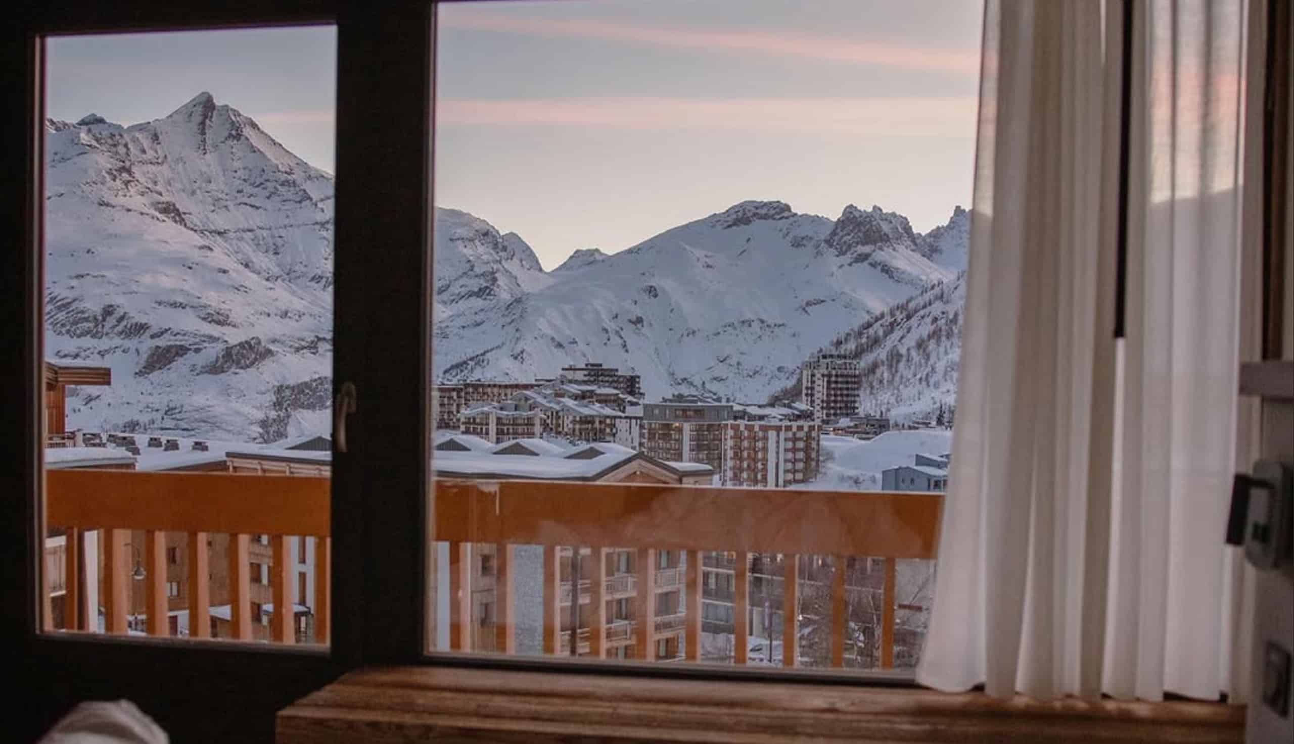 Vue d'une fenêtre sur des montagnes enneigées et une station de ski. Des sommets imposants et des bâtiments hivernaux sont visibles, avec un balcon en bois et un rideau.