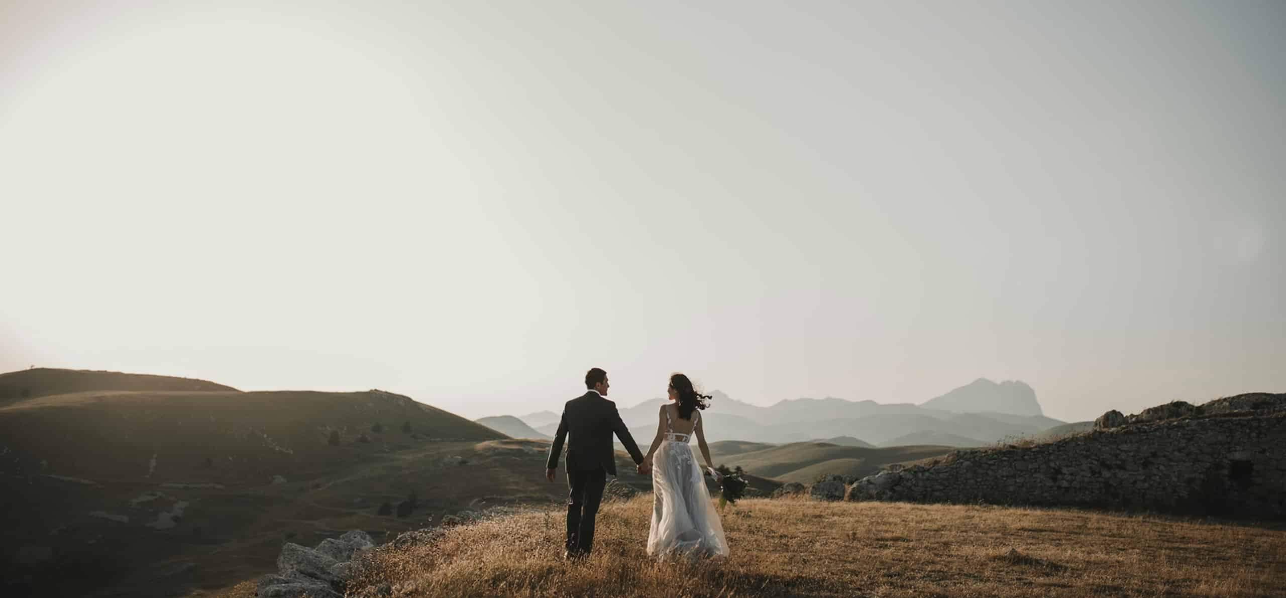 Un couple en tenue de mariés marche main dans la main sur une colline herbeuse et sèche. Des montagnes s'étendent à l'horizon sous un ciel lumineux et dégagé.