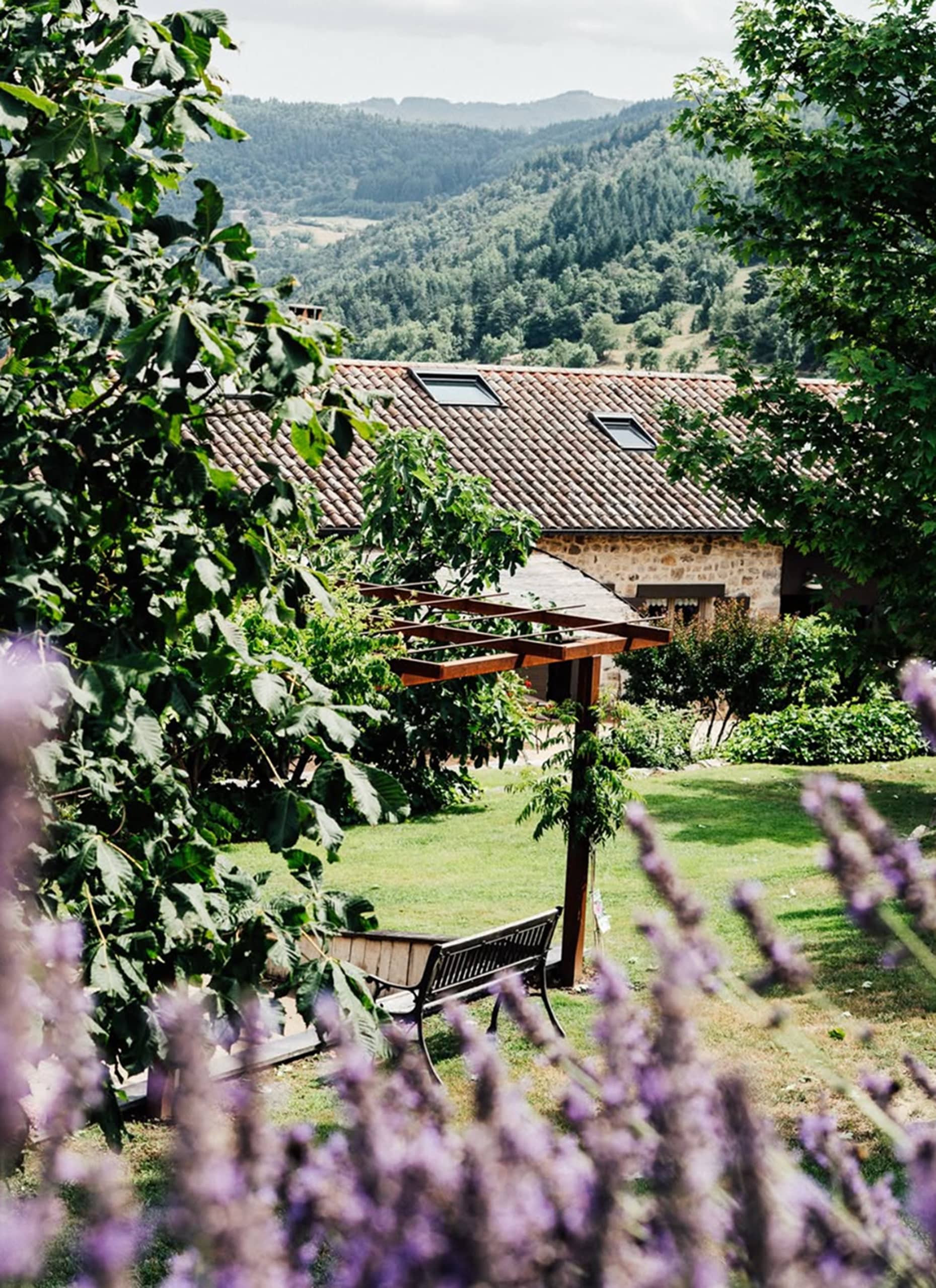Maison en pierre au toit tuilé dans un jardin verdoyant, avec pergola et banc. Lavande floue au premier plan. En arrière-plan, des montagnes boisées s'étendent sous un ciel clair.