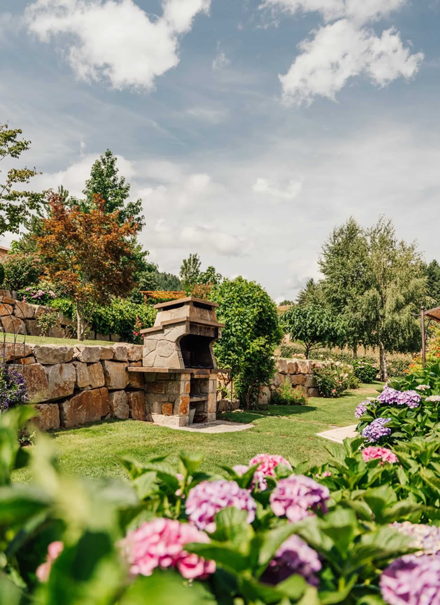Un barbecue en pierre se dresse sur une pelouse verte, entouré de murs en pierre, de parterres d'hortensias colorés et d'arbres luxuriants sous un ciel nuageux.