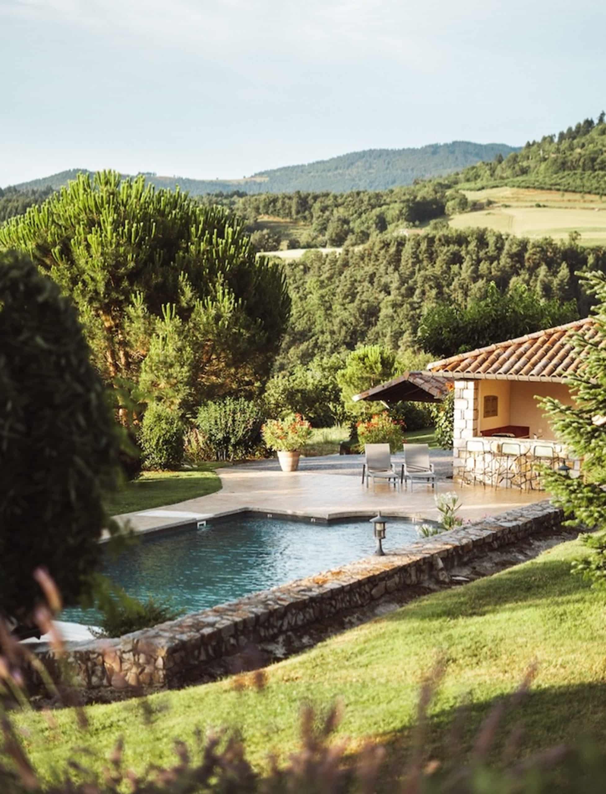 Une piscine rectangulaire et un pool house en pierre avec bar sont entourés de végétation luxuriante et de collines boisées sous un ciel clair.