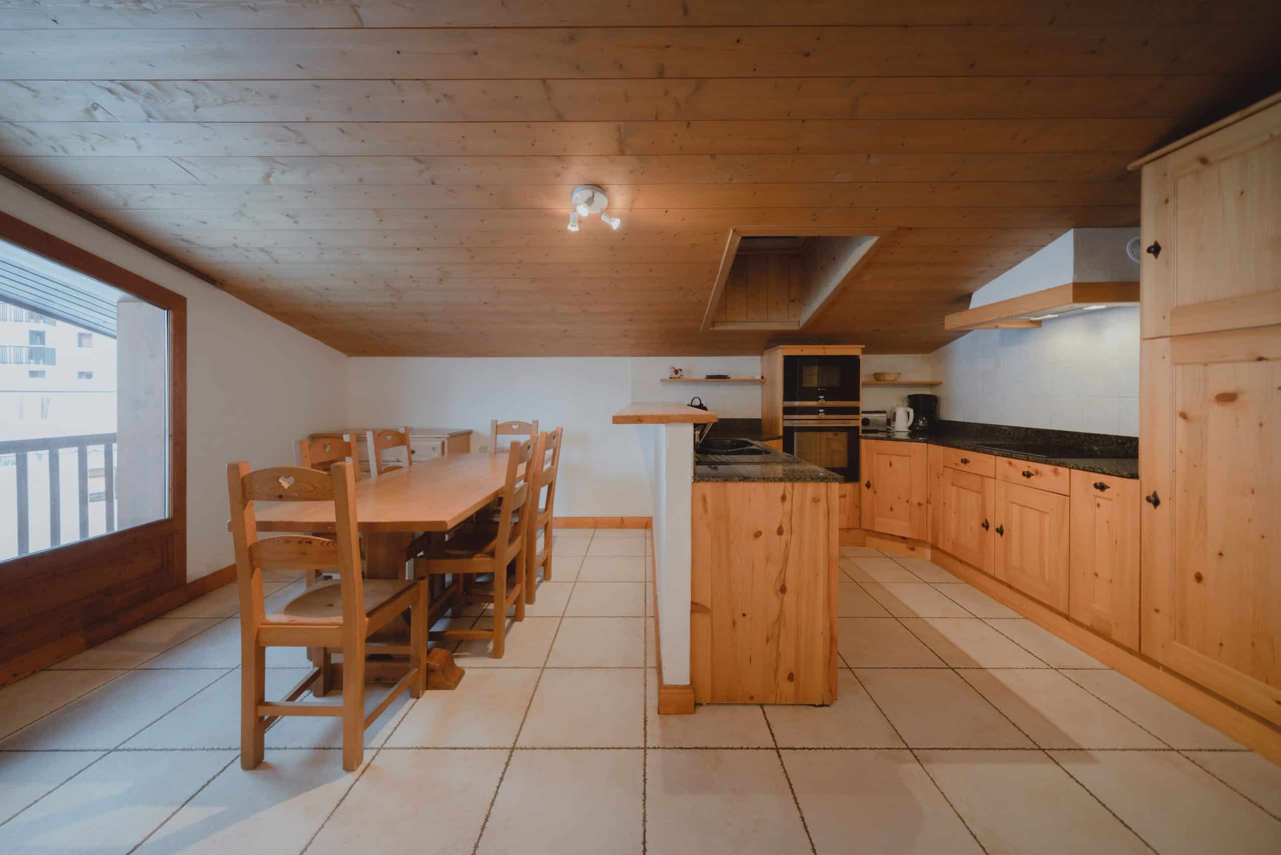 Cuisine-salle à manger spacieuse aménagée avec une longue table en bois, chaises, et des meubles de cuisine en pin clair. L'espace bénéficie d'une grande porte-fenêtre et d'un plafond lambrissé.