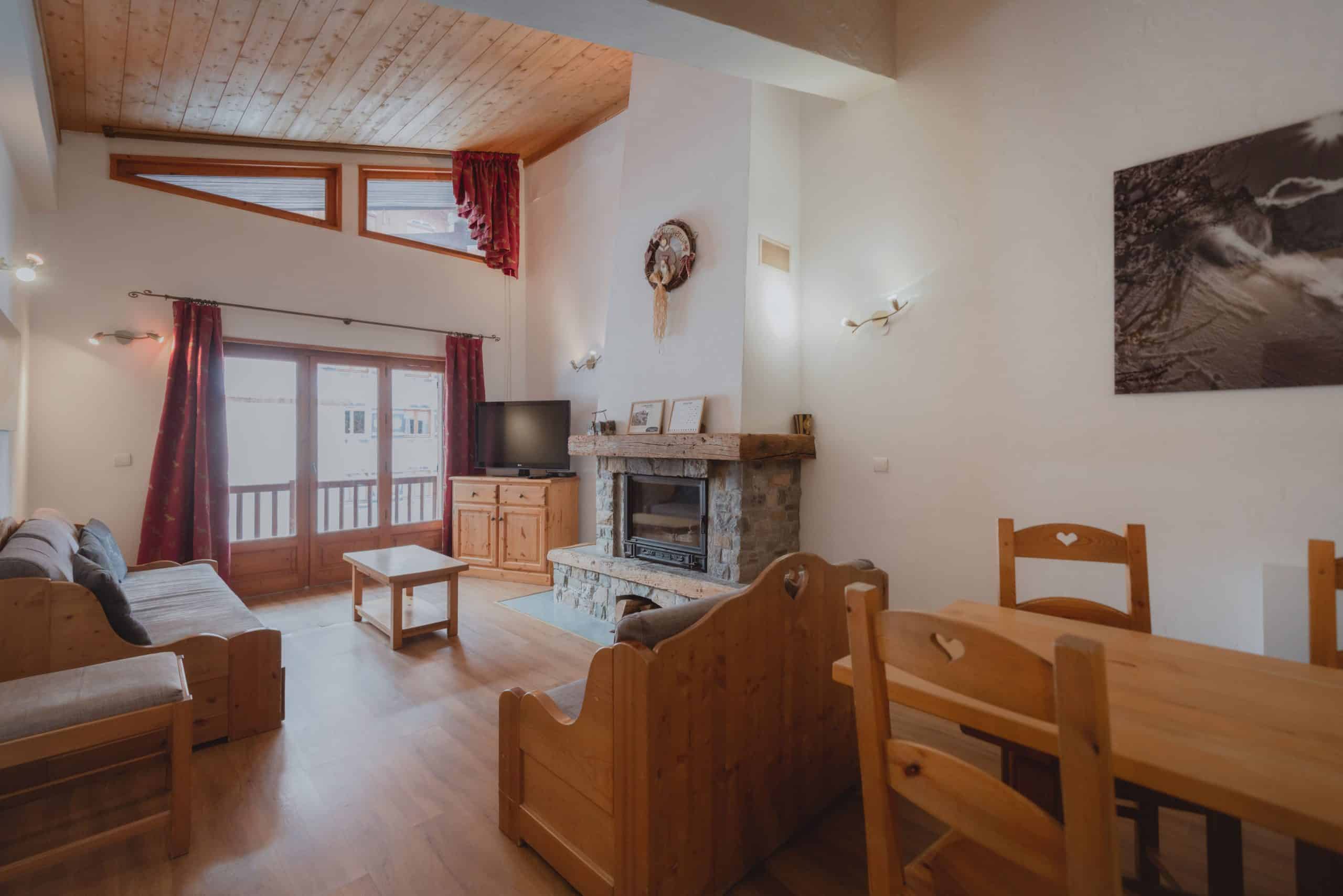 Chaleureux salon/salle à manger rustique avec mobilier en bois clair (cœurs découpés), cheminée en pierre, téléviseur, et accès à un balcon. Le plafond est lambrissé.