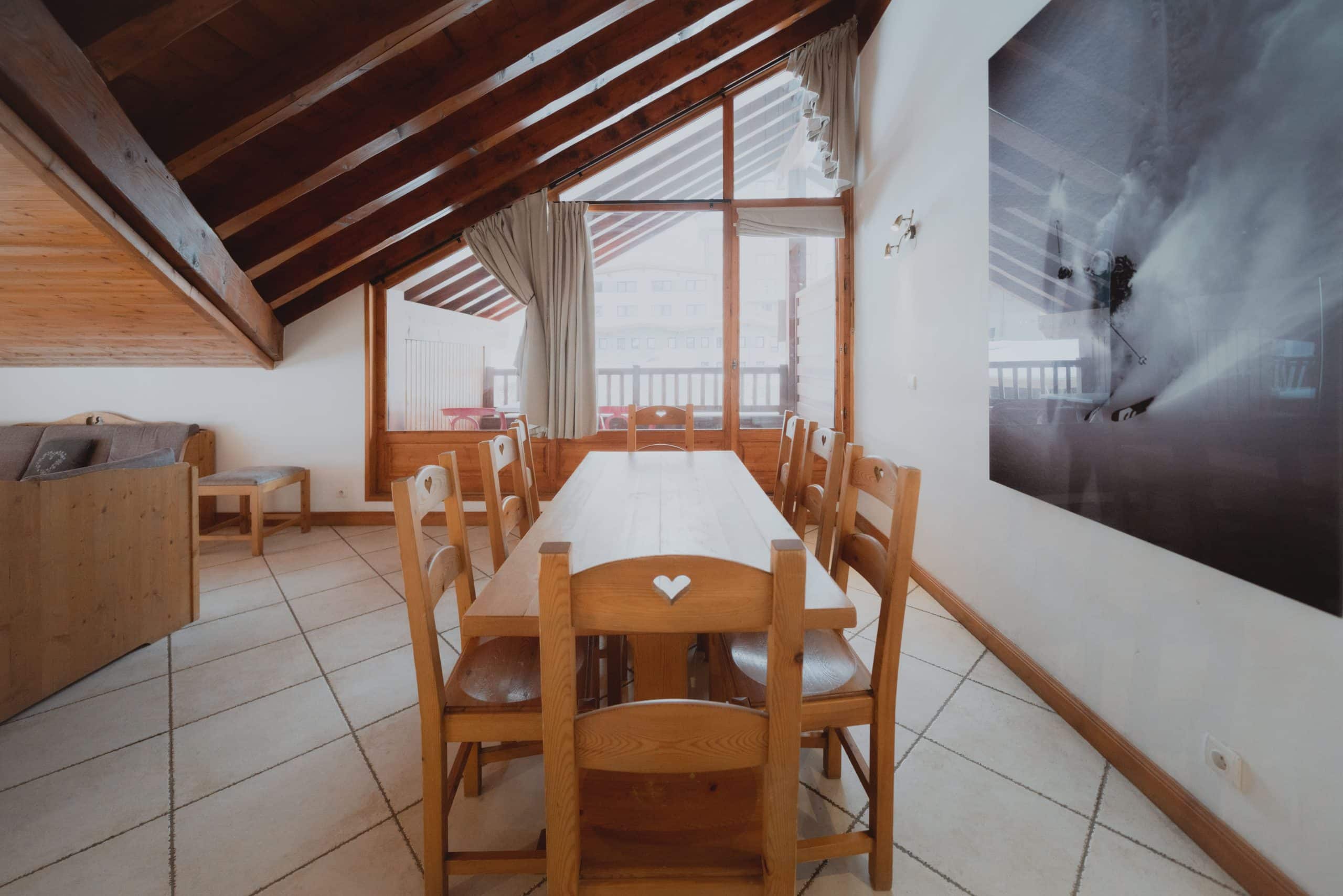 Une longue table à manger en bois clair, entourée de chaises aux cœurs sculptés. La pièce, type chalet, a un plafond à poutres, de grandes fenêtres et un tableau de skieur sur un mur blanc.