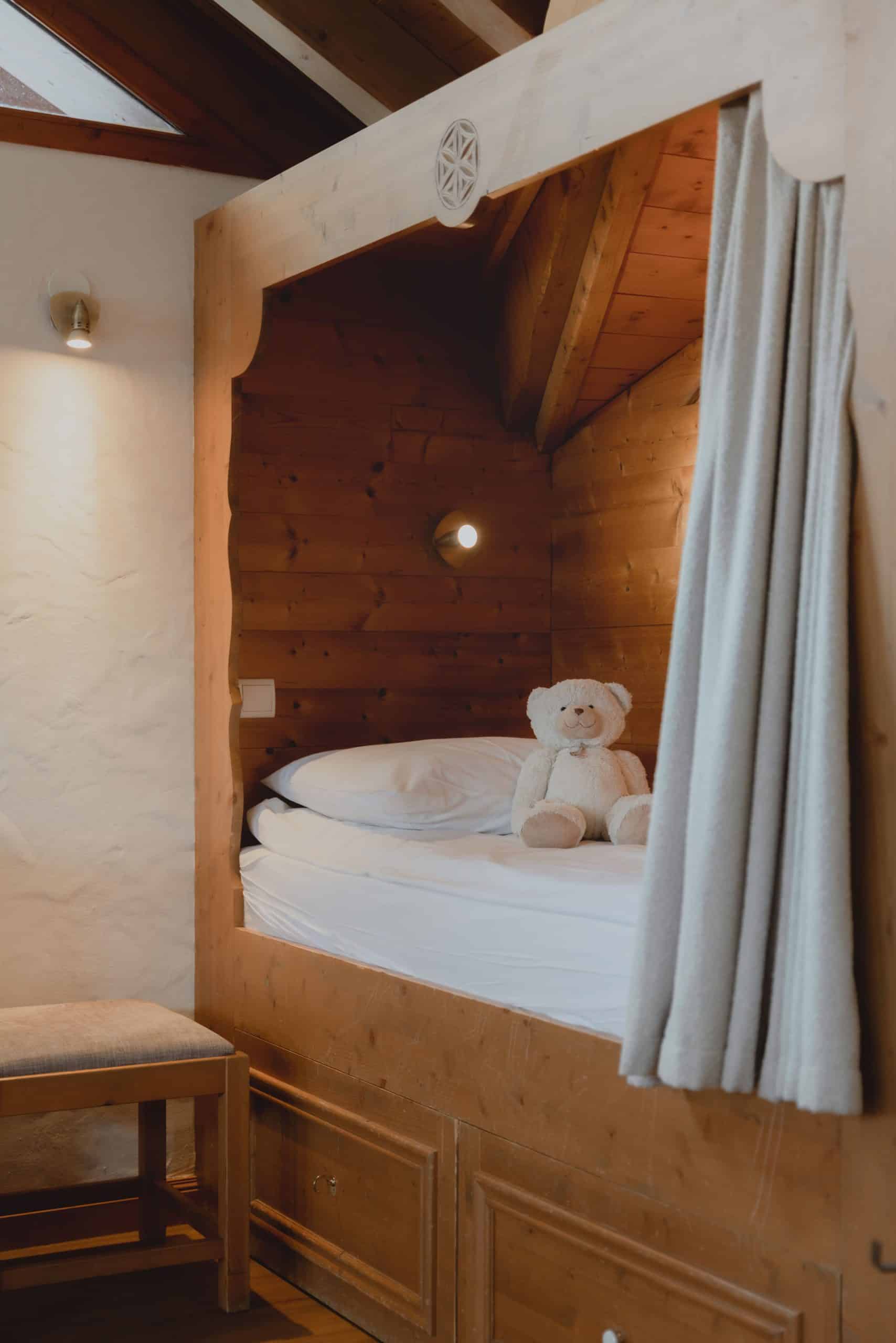 Un ourson en peluche blanc est assis sur un lit dans un lit cabine en bois lambrissé, éclairé par une applique murale et doté d'un rideau.