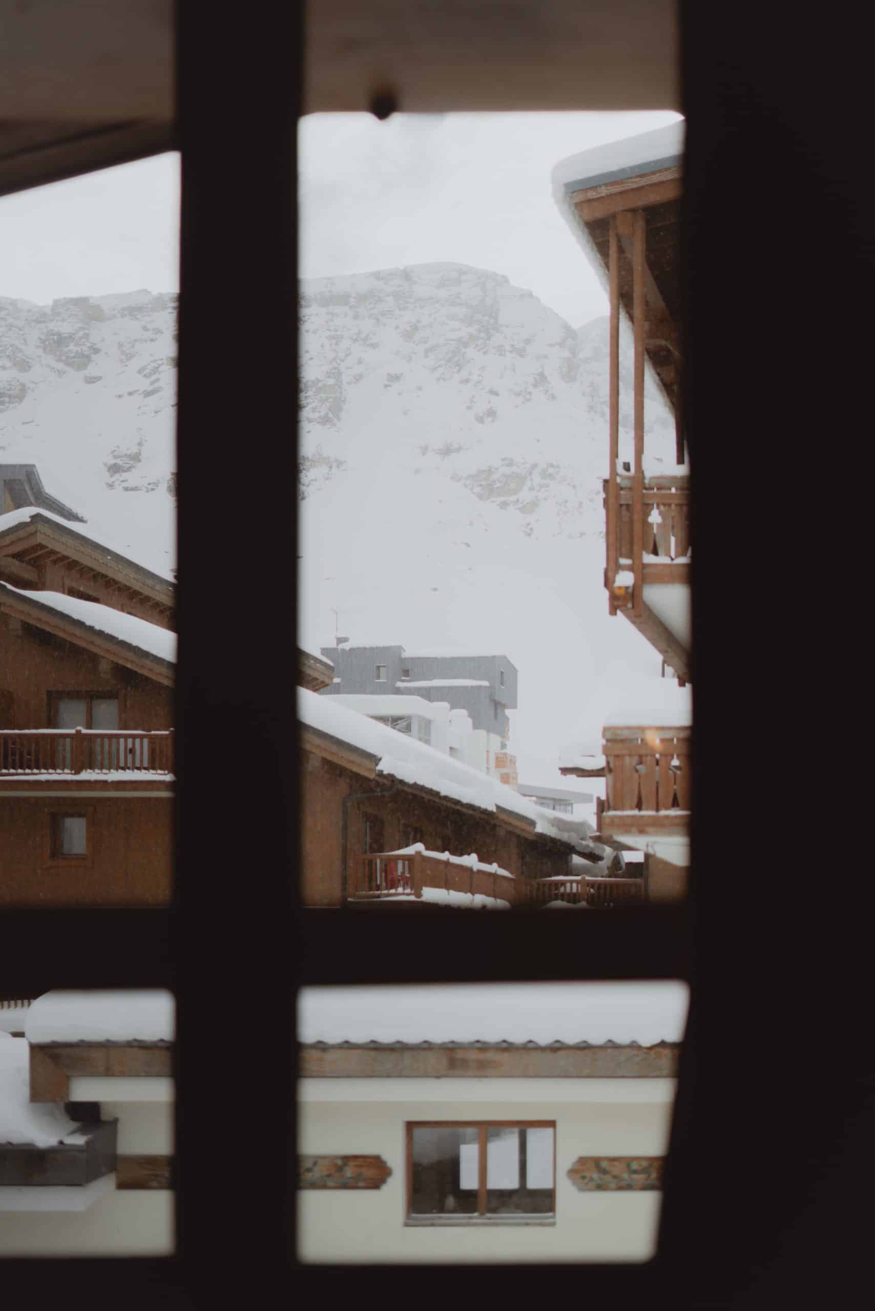 Village montagnard enneigé. Ses chalets en bois et bâtiments modernes sont recouverts de neige, tombant légèrement. Le tout est encadré par une fenêtre sombre, devant une imposante montagne blanche.