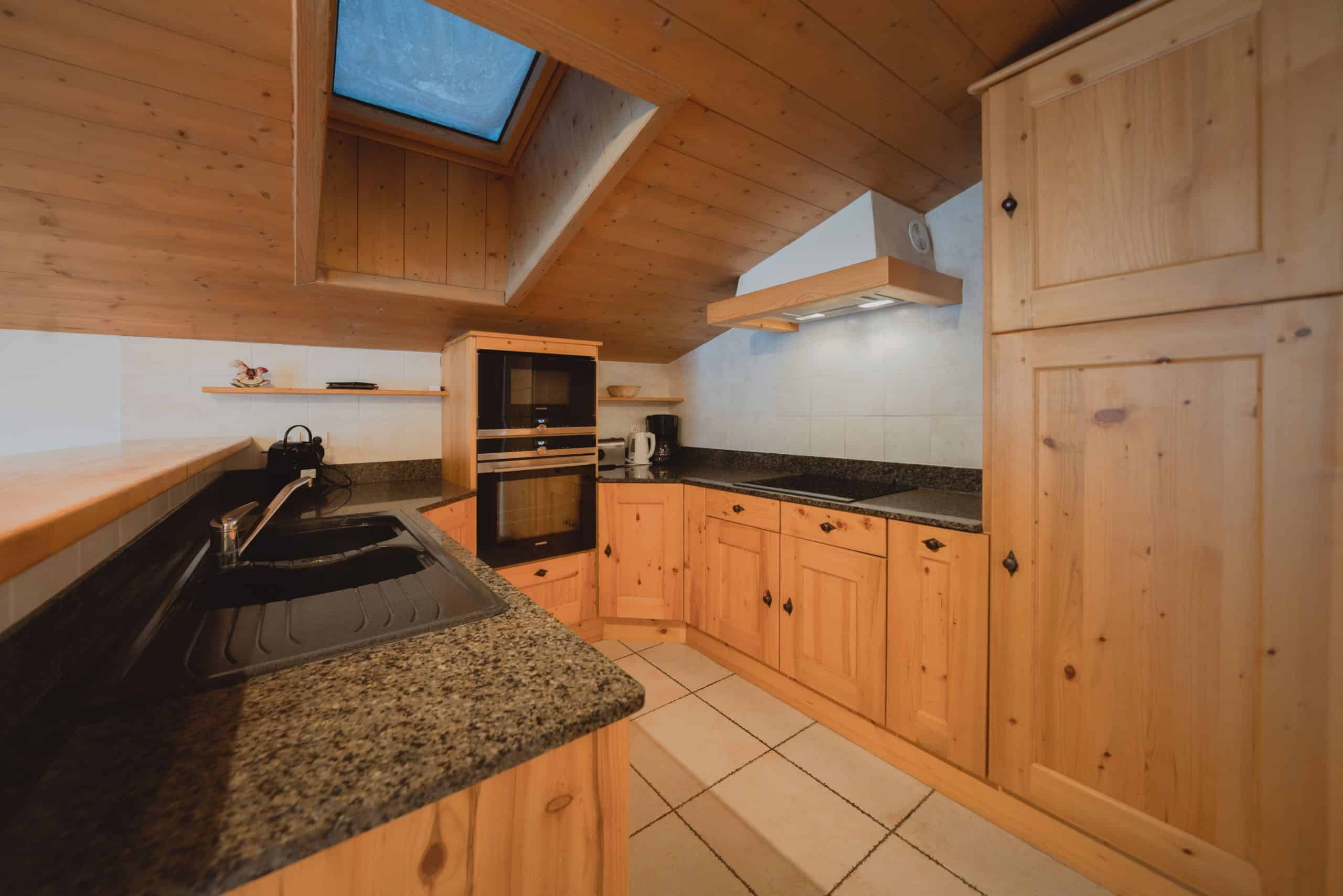 Cuisine mansardée avec meubles et plafond en bois clair. Elle comprend un évier, four, micro-ondes, plaque de cuisson, puits de lumière et sol carrelé.
