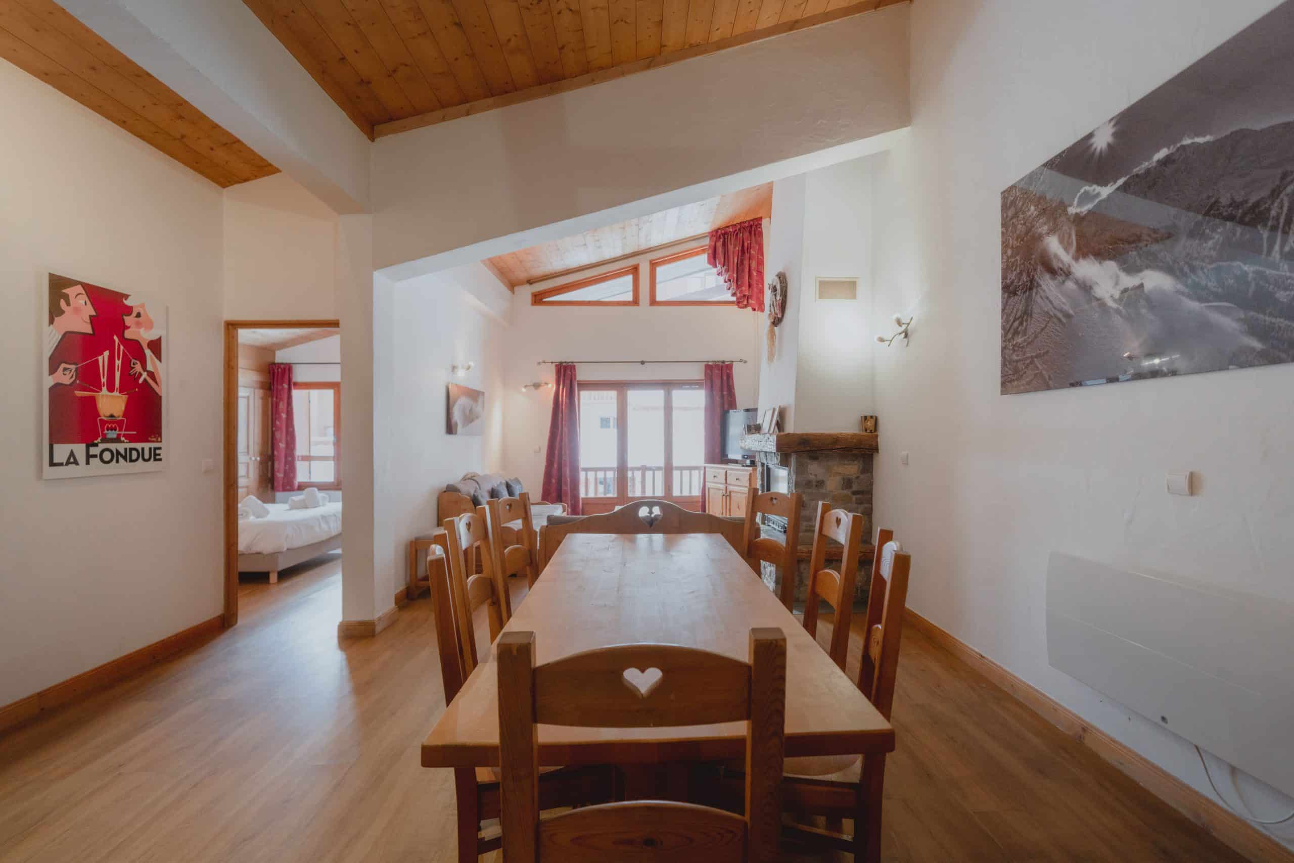 Une longue table en bois avec ses chaises occupe le centre d'une pièce lumineuse au plafond mansardé en bois. Une affiche "LA FONDUE" est sur le mur gauche, un salon avec cheminée est visible à droite.
