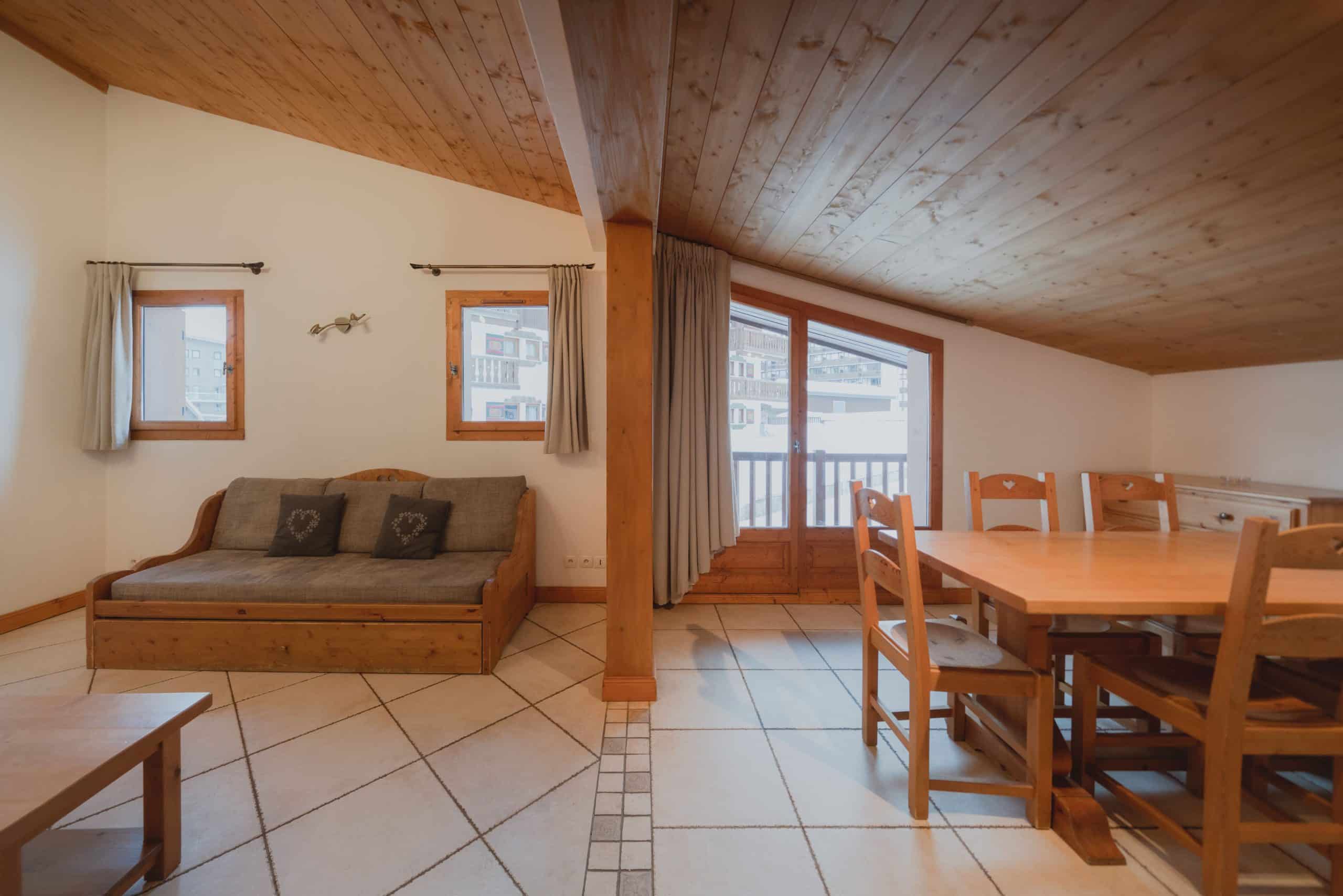 Pièce mansardée meublée d'un canapé-lit en bois et d'une table à manger avec chaises, sur carrelage clair. Plafond en bois et fenêtres donnant sur un paysage enneigé.