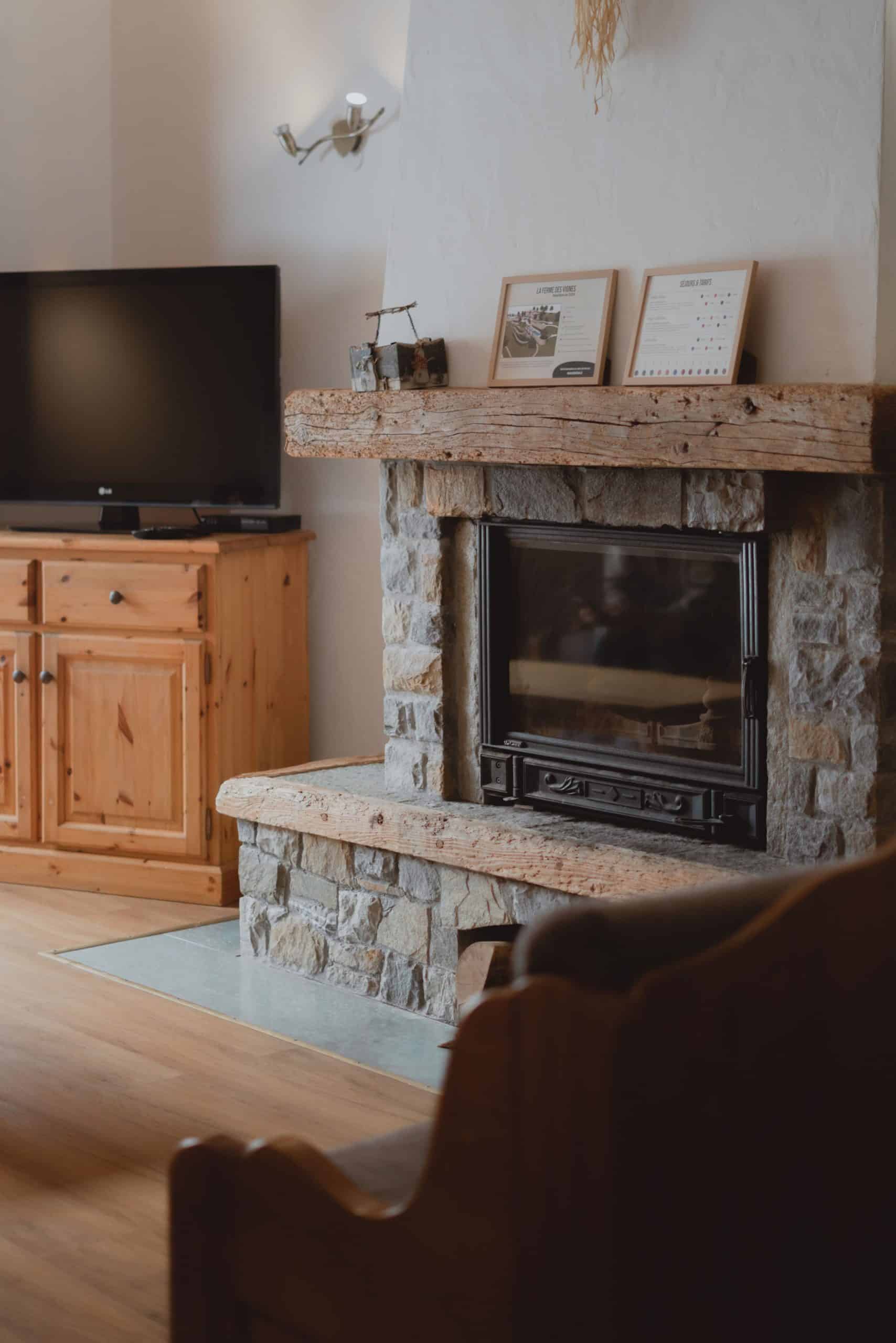 Un grand foyer en pierre surmonté d'une poutre en bois accueille deux cadres. À sa gauche, un meuble en pin porte une télévision. Texte : "LA FERME DES VIGNES Résidence de vacances - 2021" et "SÉJOURS & TARIFS".