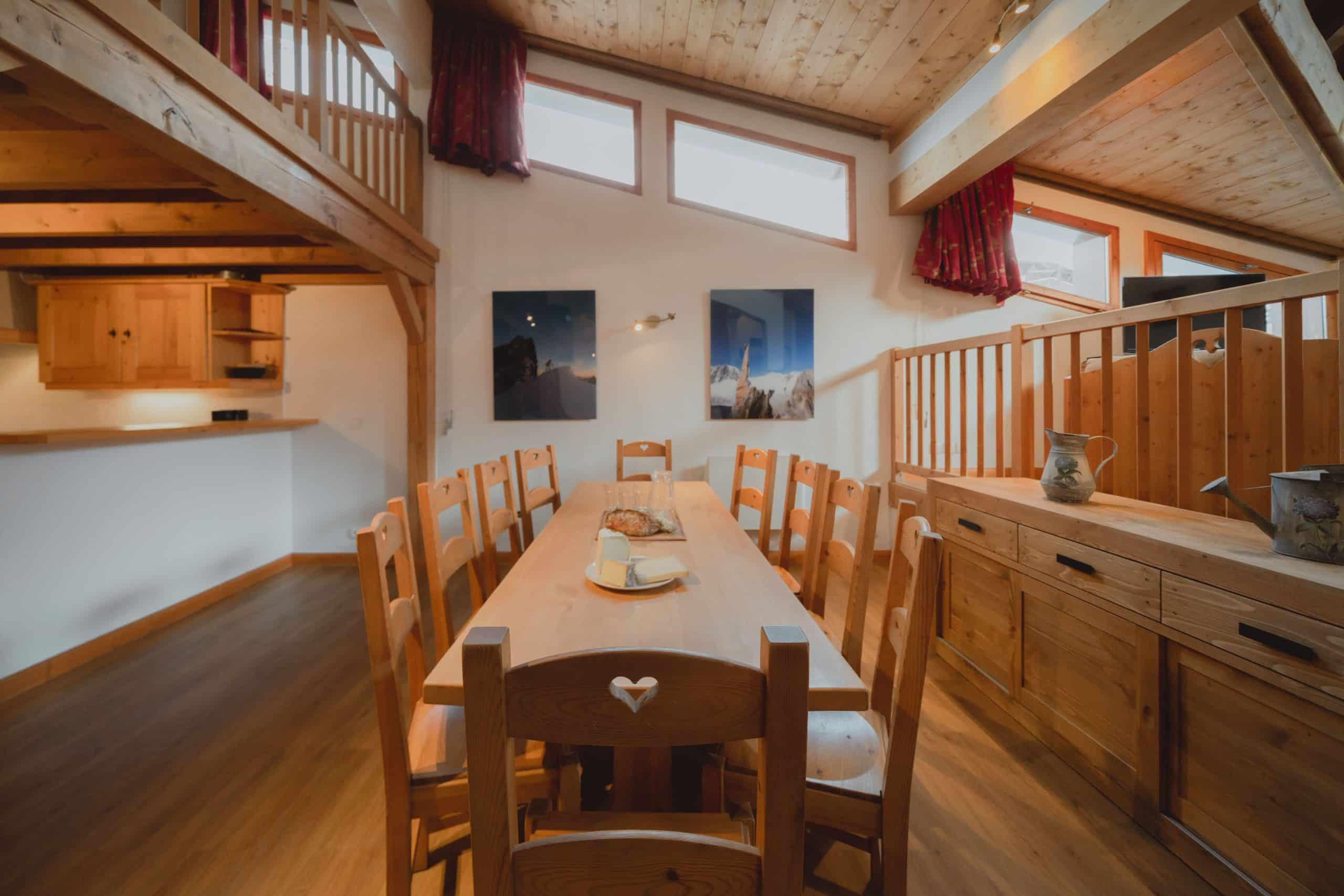 Une longue table en bois et ses chaises sont dressées avec du pain et du fromage dans une salle à manger lumineuse de style chalet, dotée d'une mezzanine et d'œuvres d'art murales de montagne.