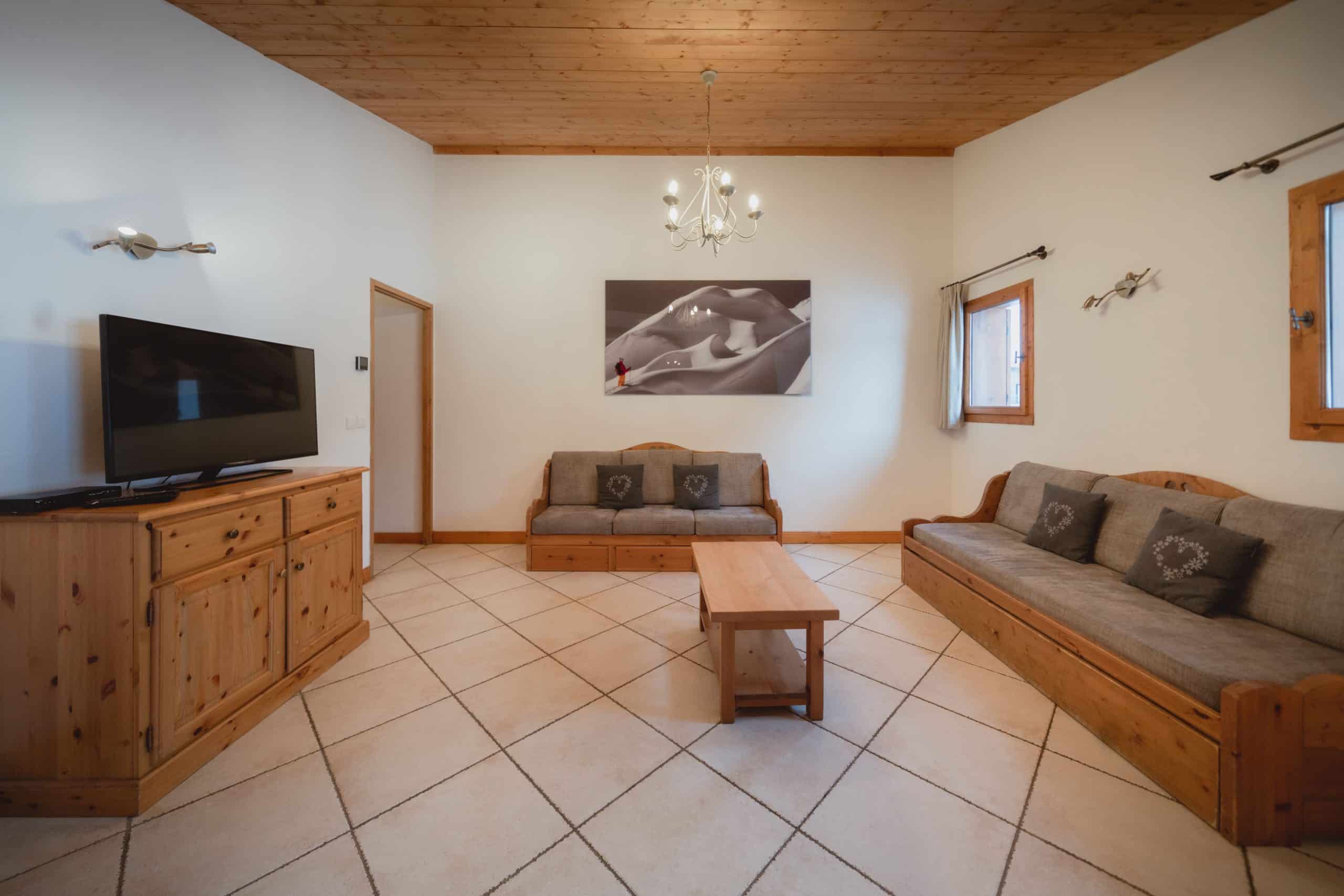 Un salon spacieux est aménagé avec deux canapés en bois, une table basse et un meuble TV. Les murs sont blancs, le plafond en bois et le sol carrelé clair.