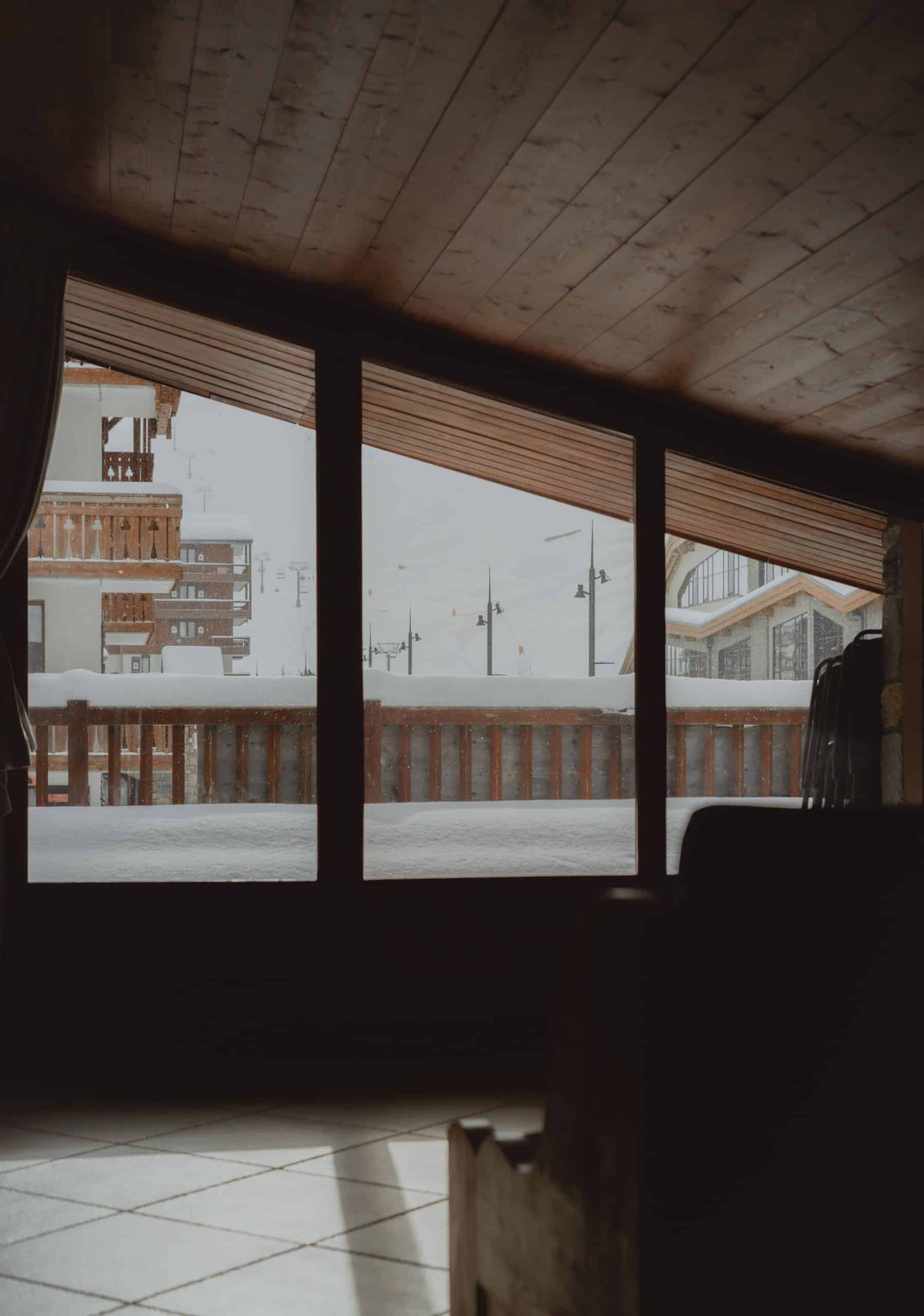 Fenêtre donnant sur une station de ski enneigée, avec bâtiments traditionnels et barrière en bois sous des flocons. L'intérieur sombre présente plafond lambrissé et sol carrelé.