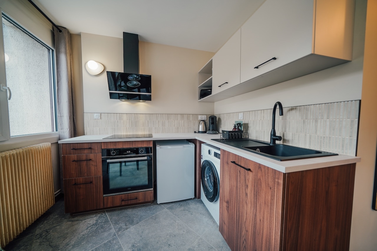Cuisine moderne : meubles bas bois, hauts clairs. Équipement incluant four "BEKO" (16:20), plaque à induction, petit frigo, lave-linge, évier noir. Fenêtre avec radiateur à gauche. Murs clairs, crédence carrelée, sol gris.