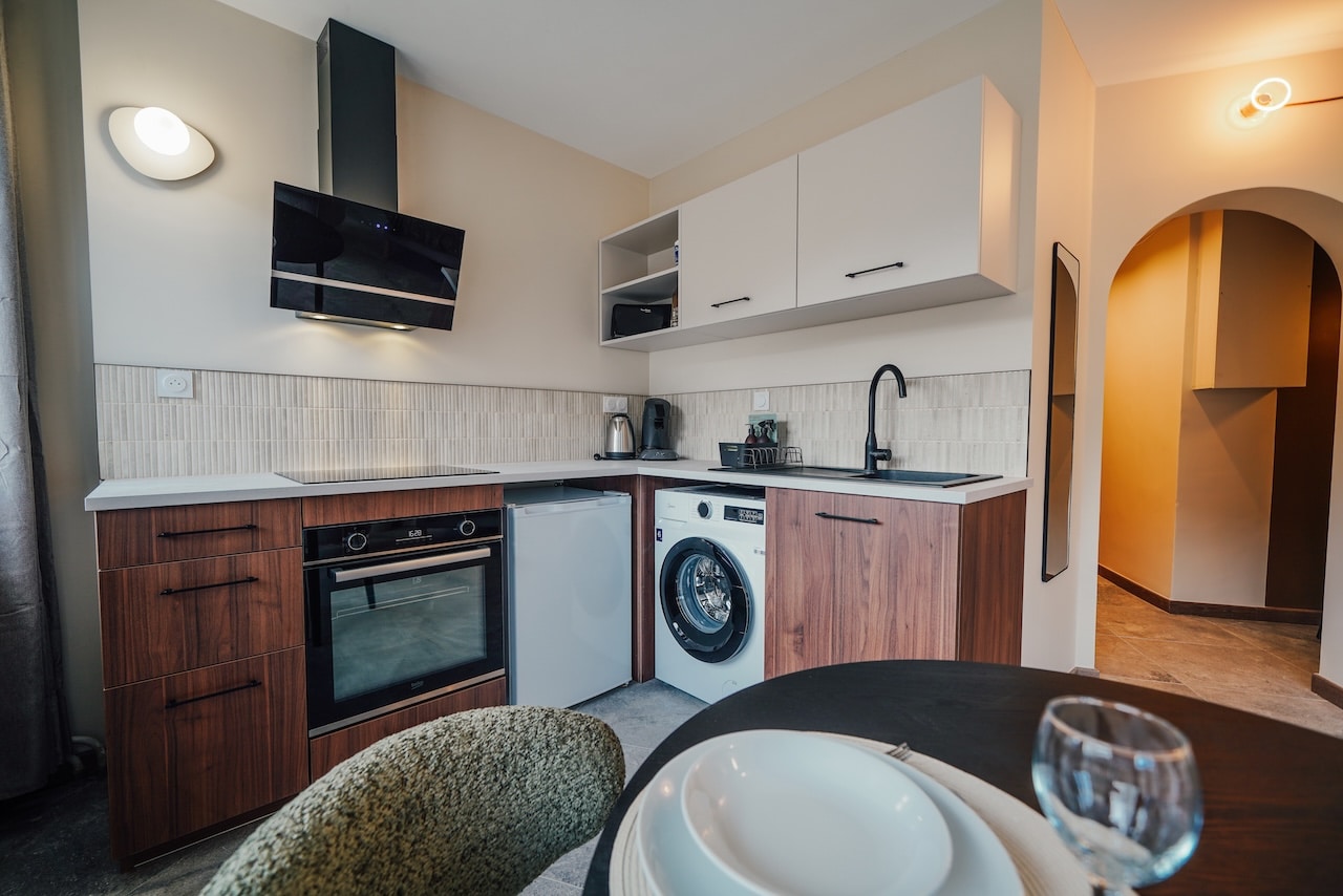 Cuisine moderne compacte avec four (BEKO, 6:20), réfrigérateur et machine à laver (10). Comptoirs clairs, meubles en bois, et crédence beige carrelée. Une table dressée est visible au premier plan, devant une arche.