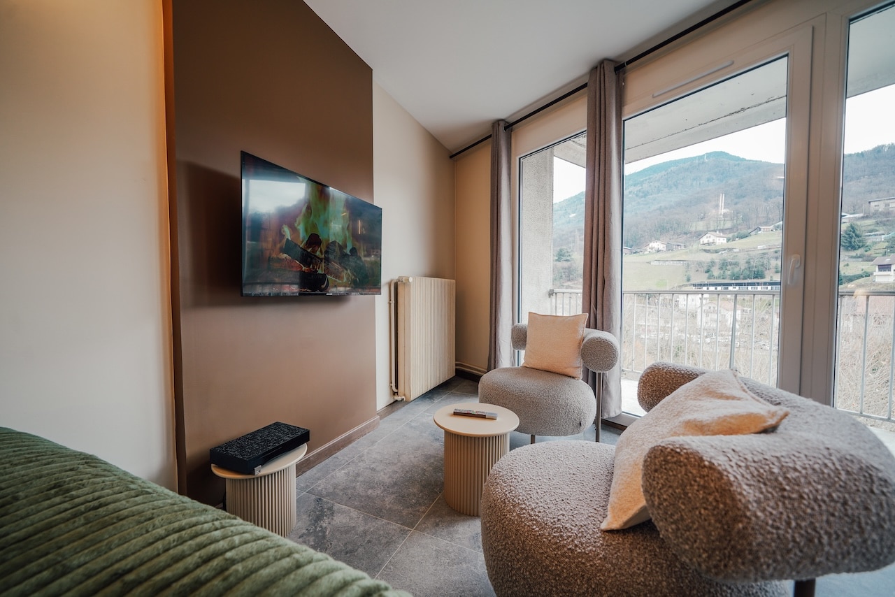 Un salon moderne avec téléviseur mural affichant un feu de cheminée. Deux fauteuils moelleux font face à des baies vitrées donnant sur un paysage montagneux avec maisons.