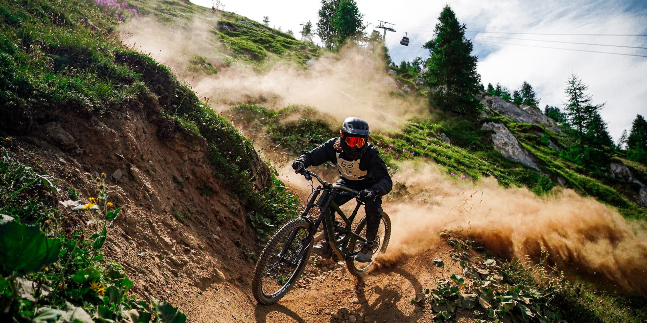 Un vététiste en maillot « FOX » et casque à lunettes rouges dévale une piste de terre sur un VTT « ALEXRIMS », soulevant un nuage de poussière sur une pente montagneuse avec télécabine.