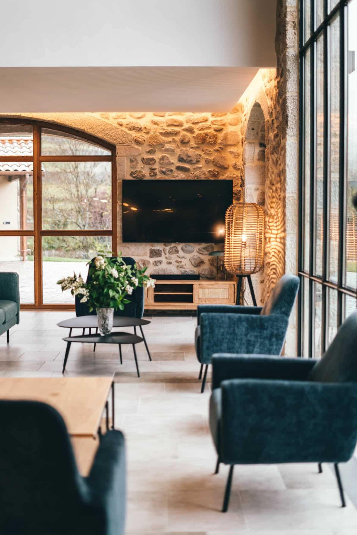 Fauteuils bleus et canapé vert meublent un salon. Une table basse fleurie est devant un mur de pierre, une TV et de grandes baies vitrées.
