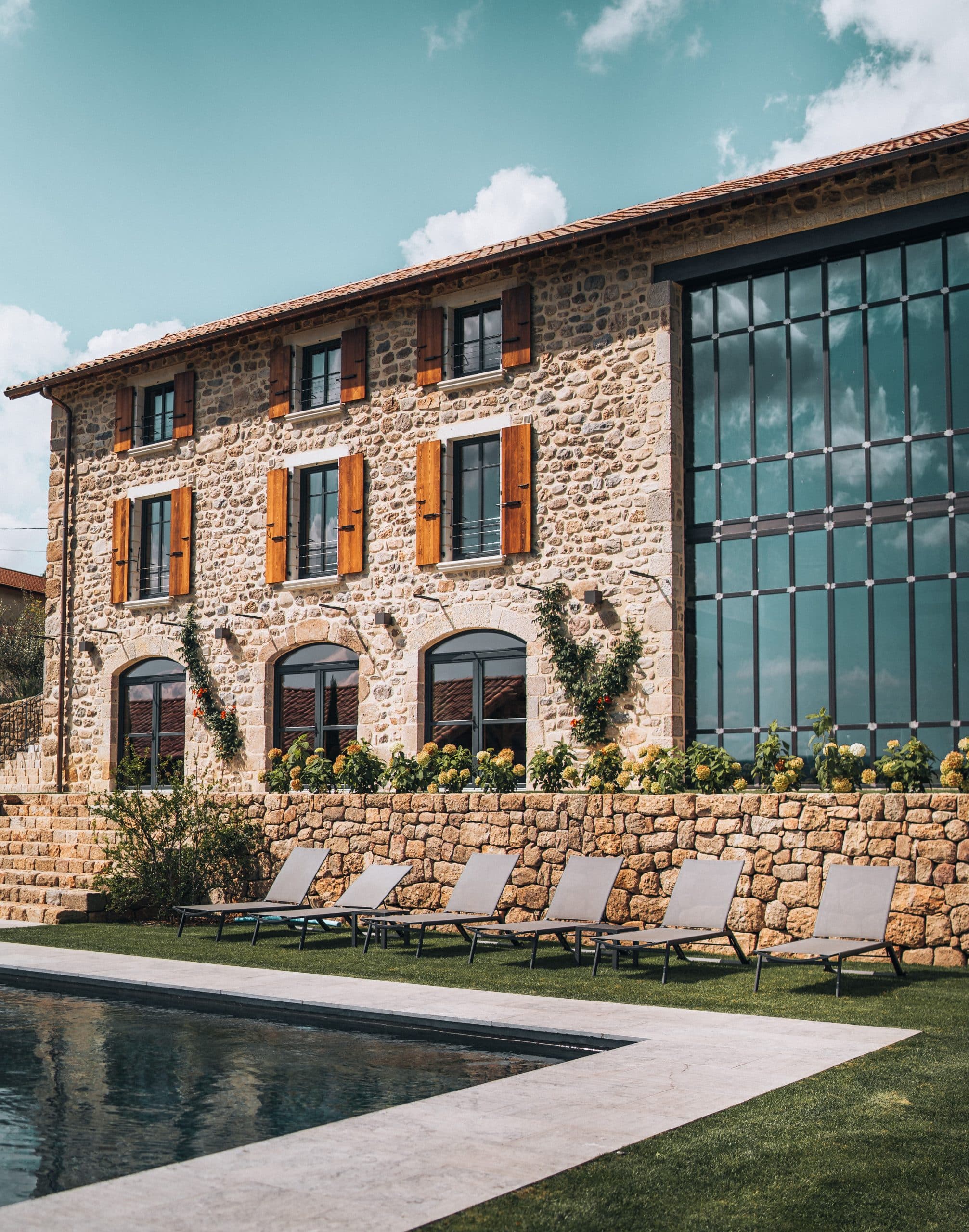 Un bâtiment en pierre avec une façade moderne vitrée se dresse au bord d'une piscine bordée de transats sur une pelouse verte, sous un ciel parsemé de nuages.