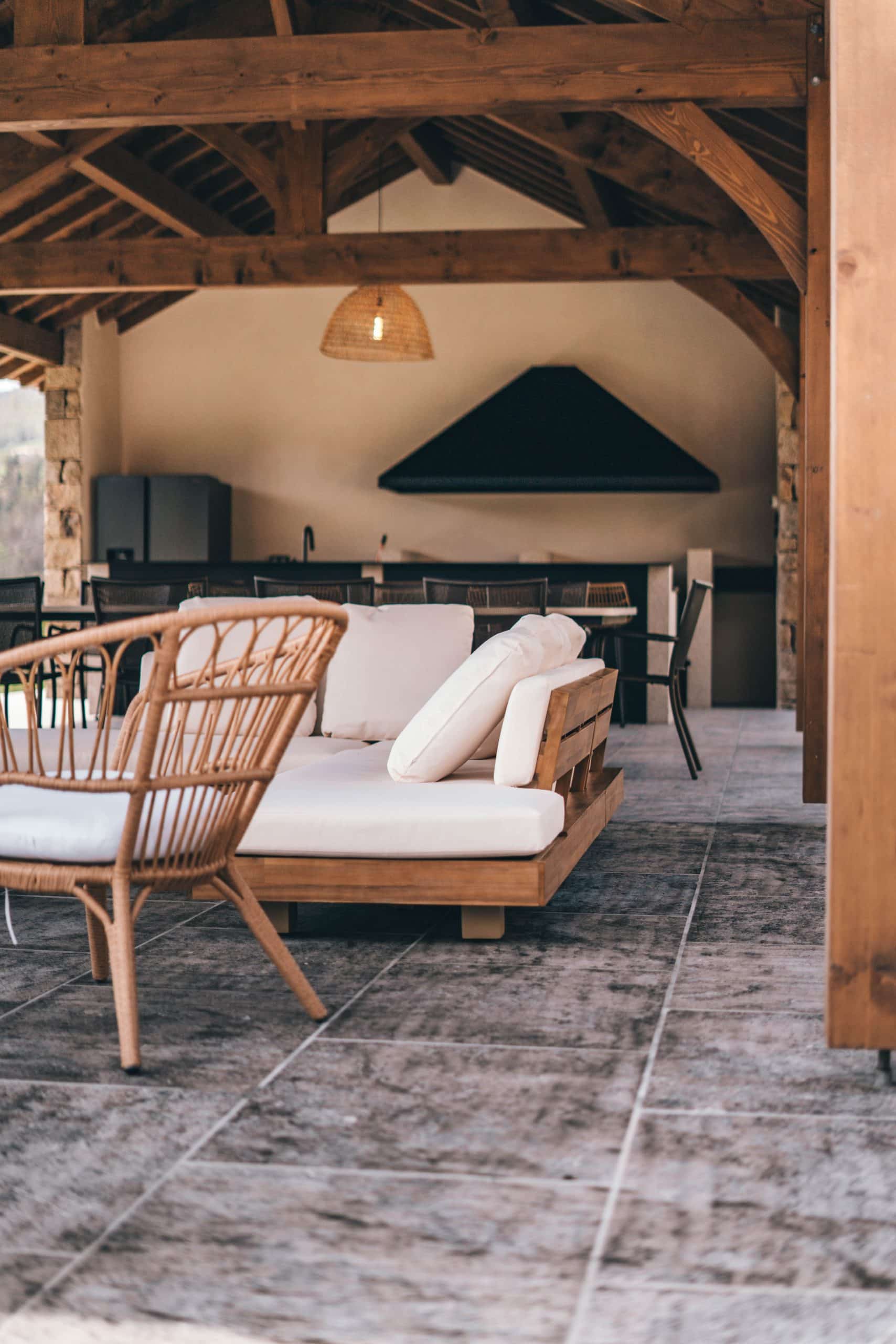 Un fauteuil en rotin et canapé-lit en bois clair à coussins blancs meublent une pergola. Au fond, une cuisine d'extérieur avec hotte noire surplombe un sol carrelé gris.