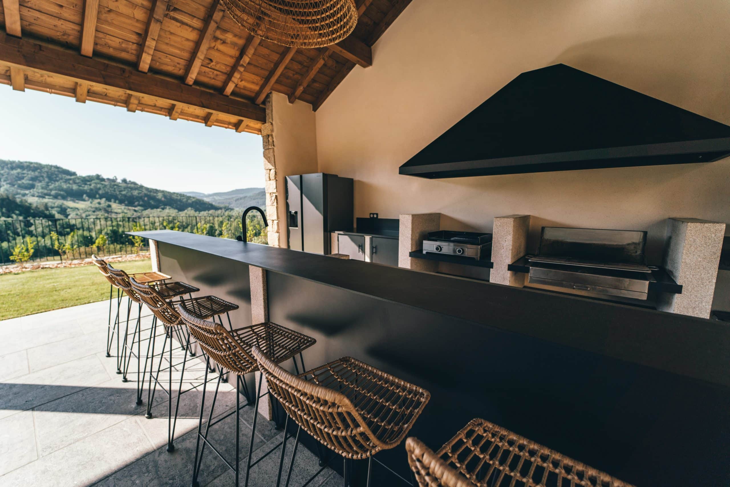 Cuisine d'été moderne sous abri en bois, avec comptoir noir, tabourets et appareils de cuisson intégrés. Elle offre une vue panoramique sur des collines verdoyantes.