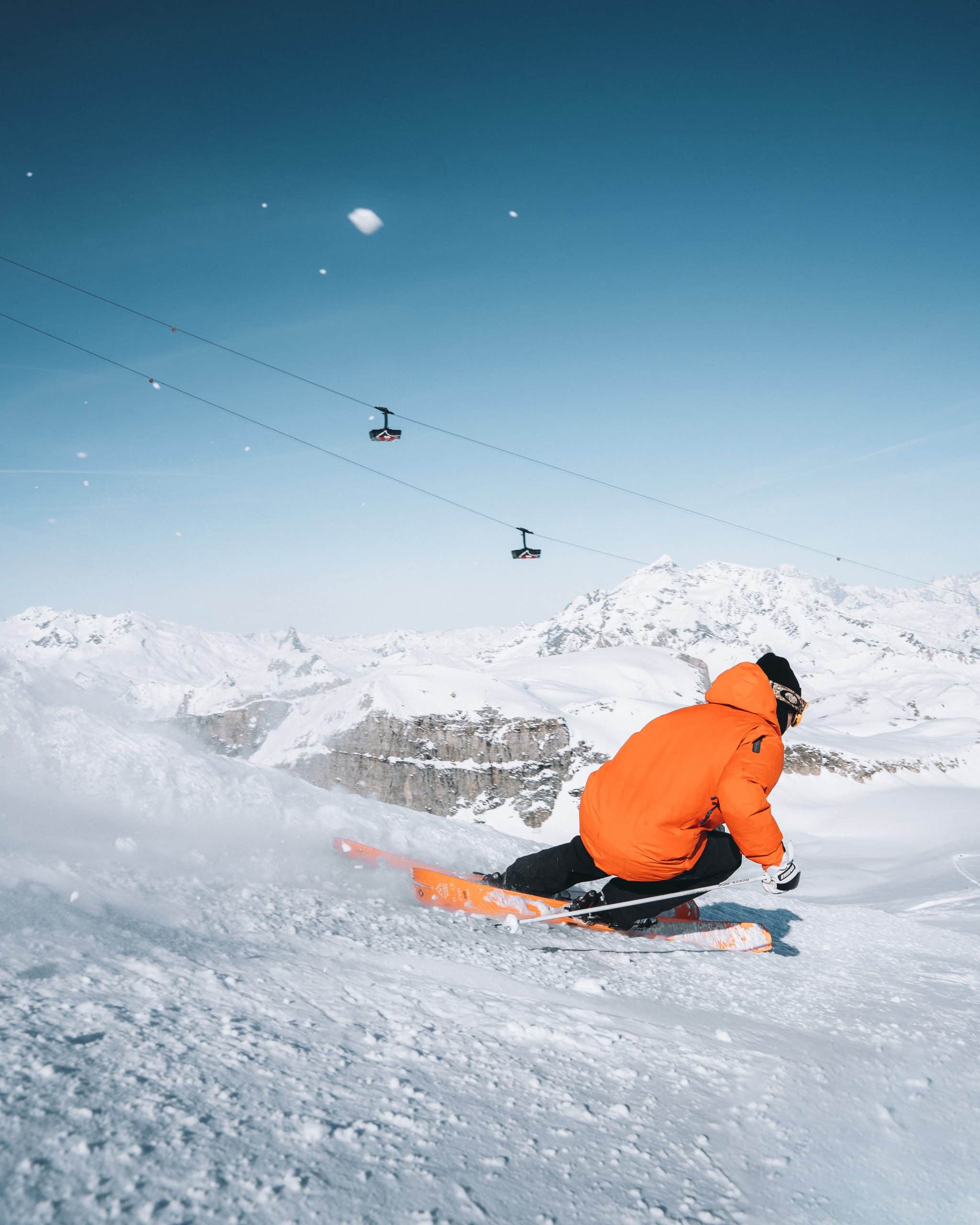 Un skieur en veste orange dévale une piste enneigée, projetant de la poudreuse. Montagnes et télécabines en arrière-plan sous un ciel bleu. Texte visible : Skis K2 RECKONER 112 FREERIDE. Bâtons MOUILLE RIDGE.