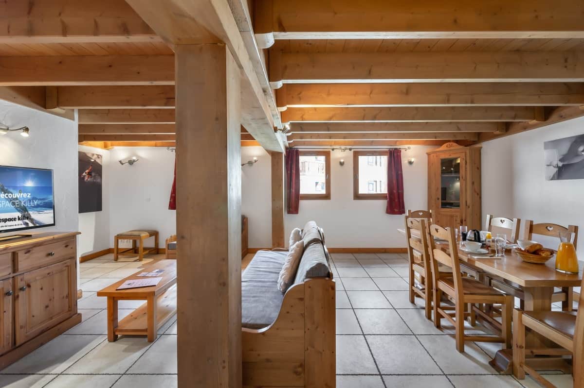 Un grand salon-salle à manger rustique avec poutres apparentes et sol carrelé clair. Une télévision affiche "découvrez SPACE KILLY espace killy". Une table en bois est dressée pour le petit-déjeuner.