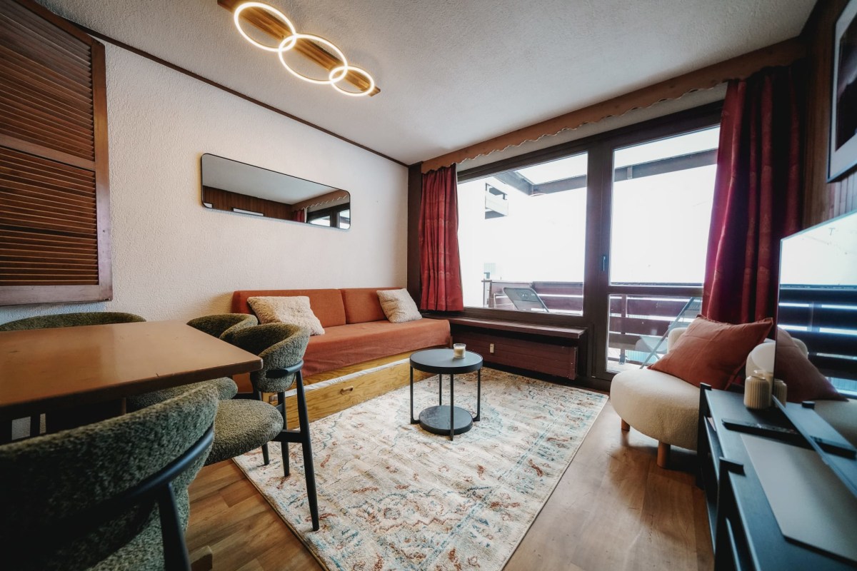 Salon moderne avec table et chaises, canapé-lit et fauteuil sur tapis clair. La pièce lumineuse s'ouvre sur un balcon extérieur. Éclairage design au plafond.