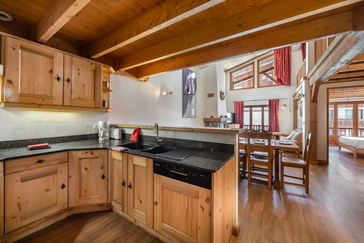 Une cuisine en bois clair s'ouvre sur une salle à manger et un salon. Le vaste intérieur, avec poutres apparentes et parquet, dégage une atmosphère chaleureuse de chalet.