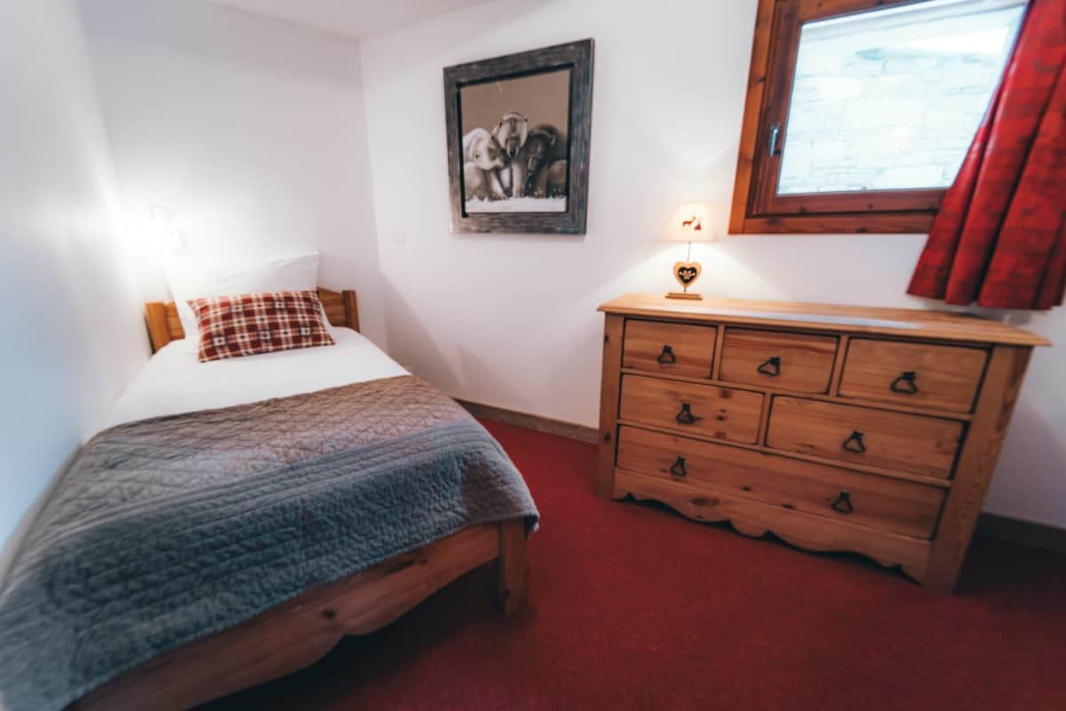 Lit simple en bois avec couette grise et coussin à carreaux rouges, dans une chambre aux murs blancs. Une commode en bois, une lampe et un tableau d'ours complètent l'espace avec moquette rouge.