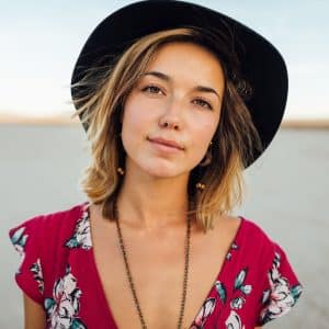 Une jeune femme, portant un chapeau noir et un top floral rouge, regarde le spectateur avec un léger sourire, sur fond de paysage extérieur clair et estompé.