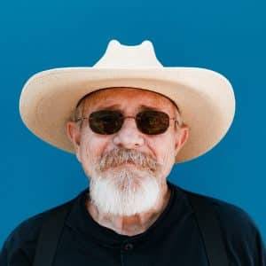 Un homme barbu aux cheveux blancs, coiffé d'un chapeau de cow-boy crème et de lunettes de soleil, regarde de face sur un fond bleu uni.