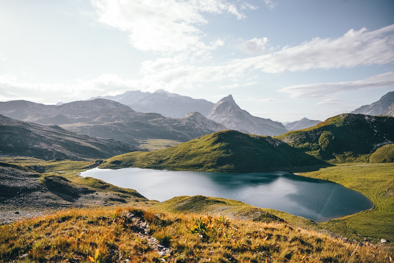 Un lac de montagne bleu foncé s'étend, entouré de collines verdoyantes et de sommets rocheux lointains, sous un ciel lumineux et nuageux, avec de l'herbe sèche au premier plan.